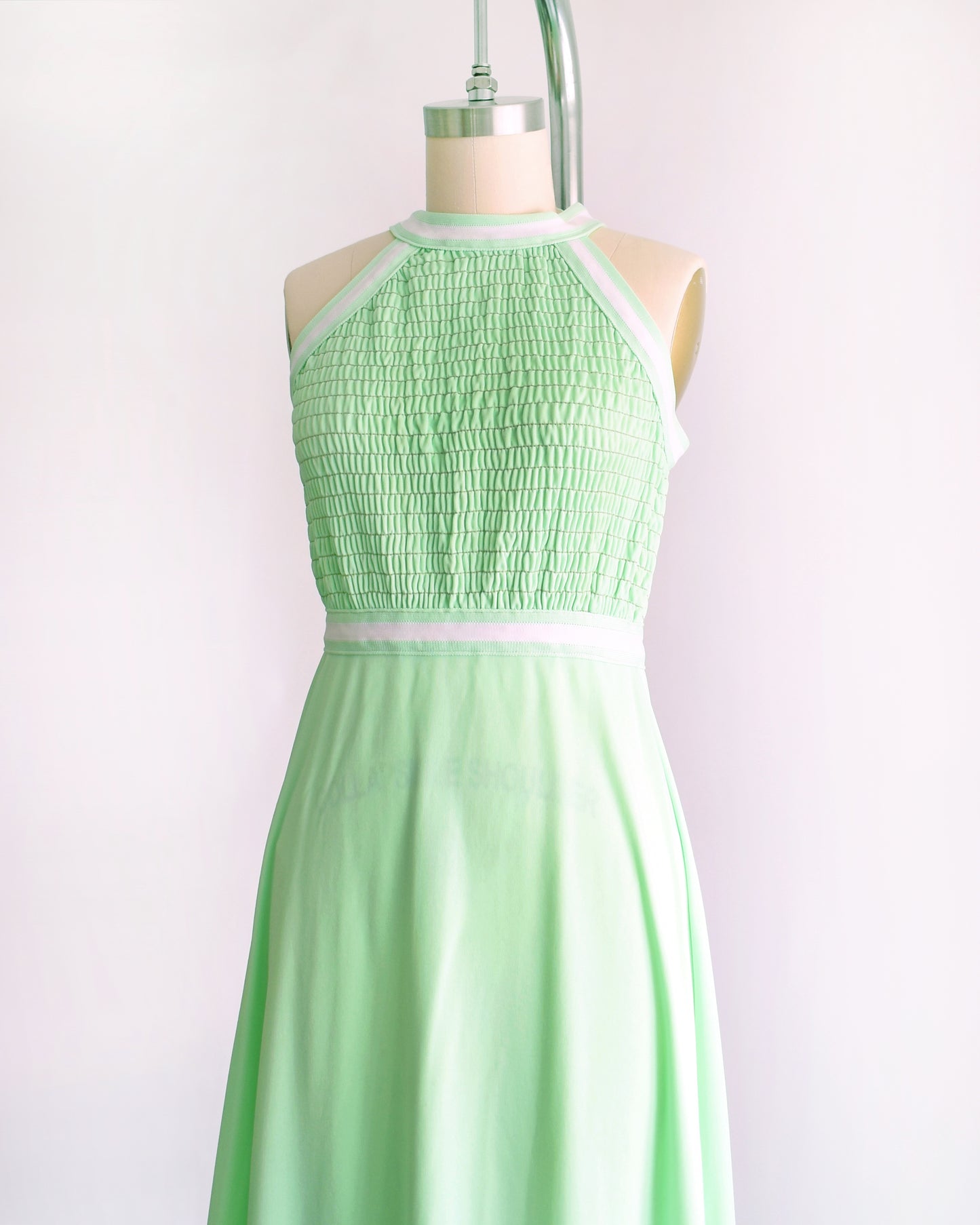 side front view of a vintage 1970s mint green sleeveless halter maxi dress with a smocked bodice and a flared skirt, displayed on a dress form against a plain light background.