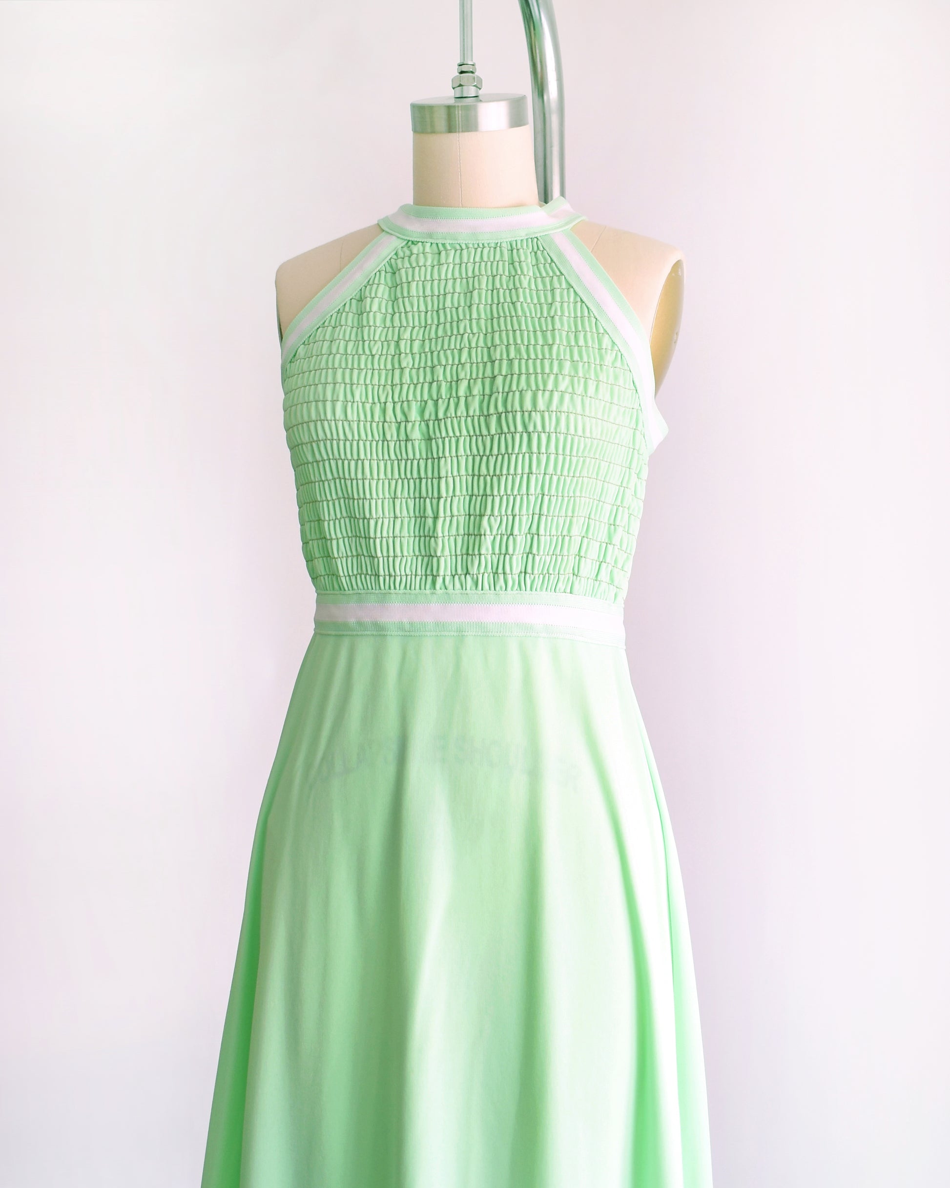 side front view of a vintage 1970s mint green sleeveless halter maxi dress with a smocked bodice and a flared skirt, displayed on a dress form against a plain light background.
