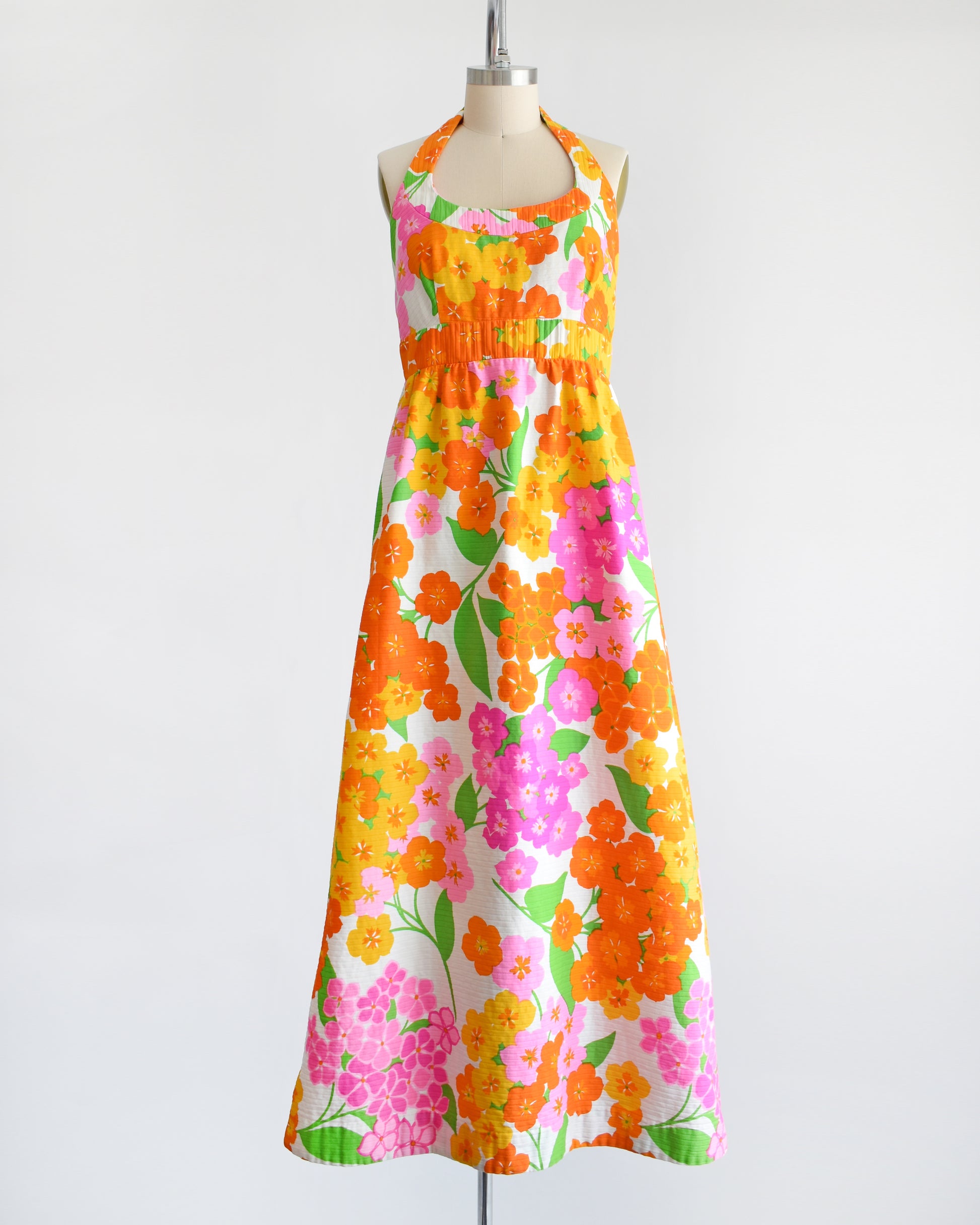 a vintage 1970s Hawaiian floral maxi dress by Malia. The dress is made from white ribbed cotton and has a large pink, yellow, orange, and green floral print covering the dress.