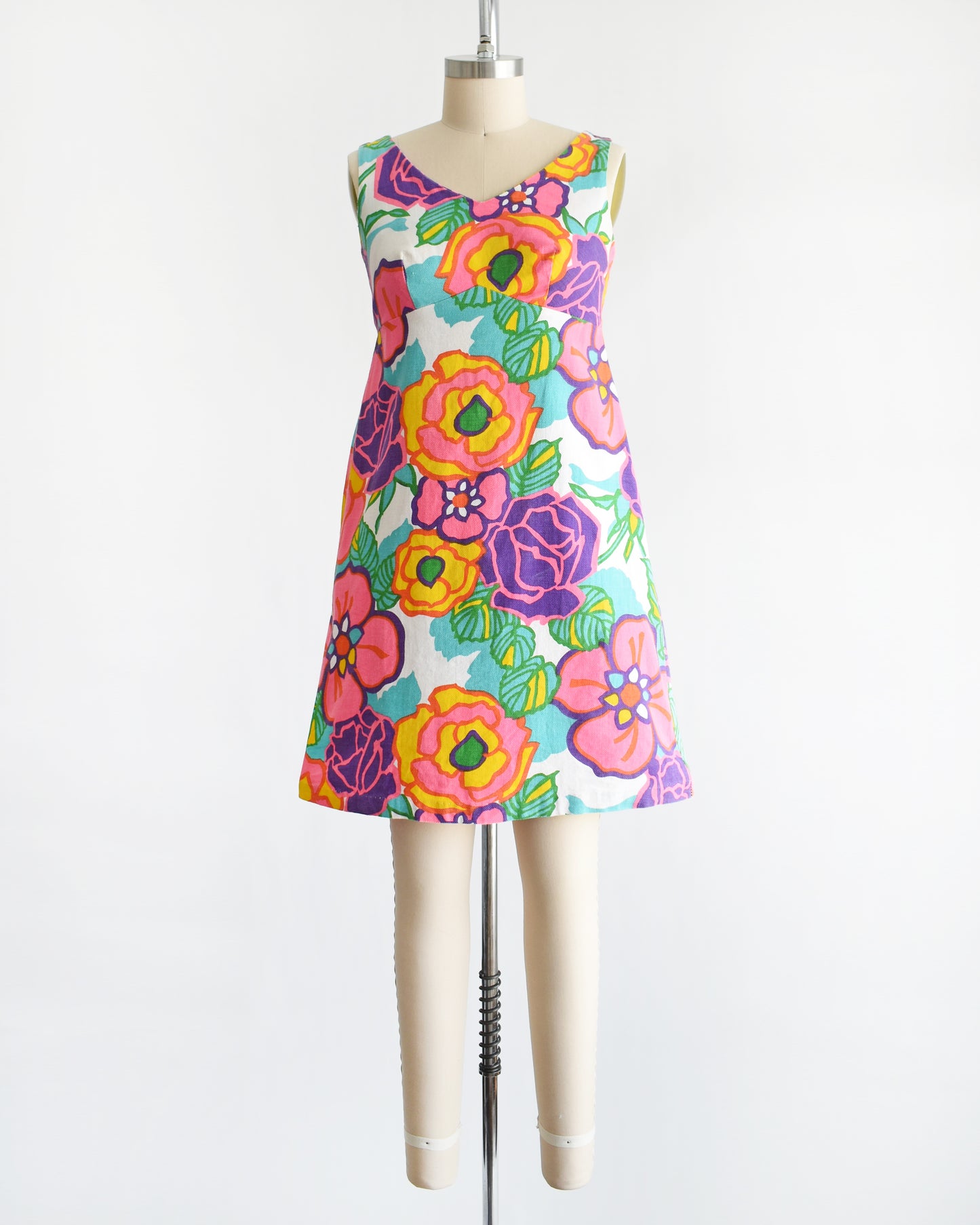 A vintage 1960s sleeveless A-line dress with a vibrant retro floral print, displayed on a cream-colored dress form mannequin against a plain white background.