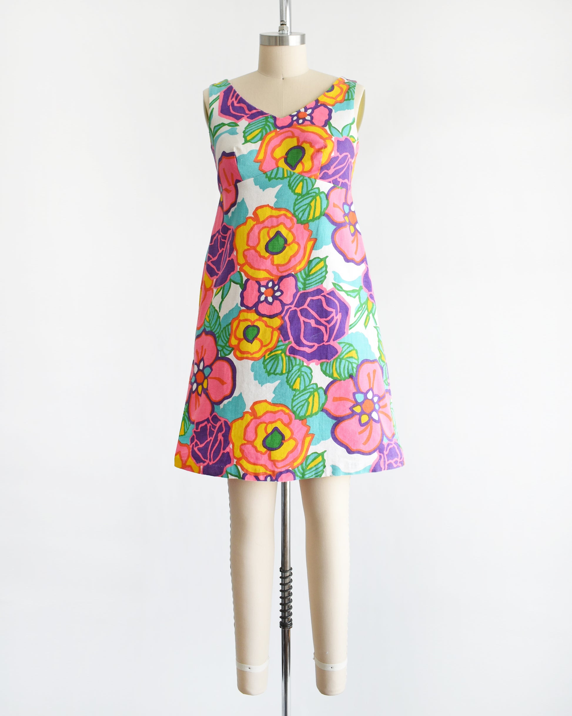 A vintage 1960s sleeveless A-line dress with a vibrant retro floral print, displayed on a cream-colored dress form mannequin against a plain white background.