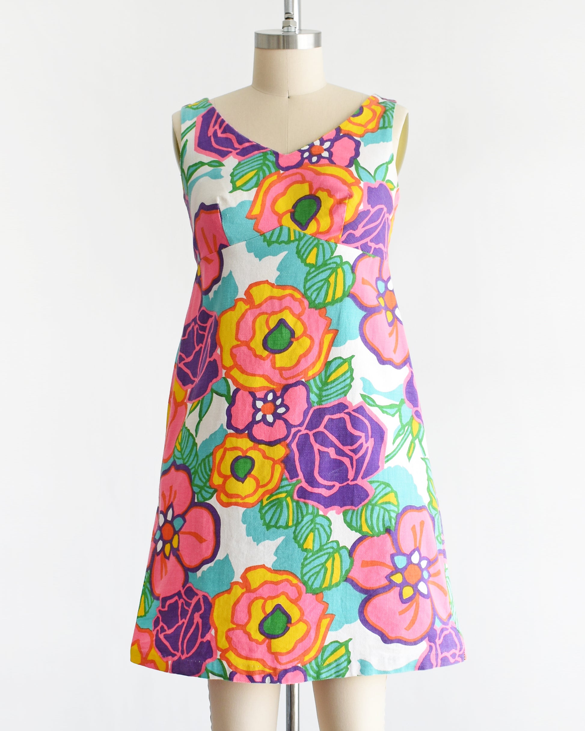 A vintage 1960s sleeveless A-line dress with a vibrant retro floral print, displayed on a cream-colored dress form mannequin against a plain white background.