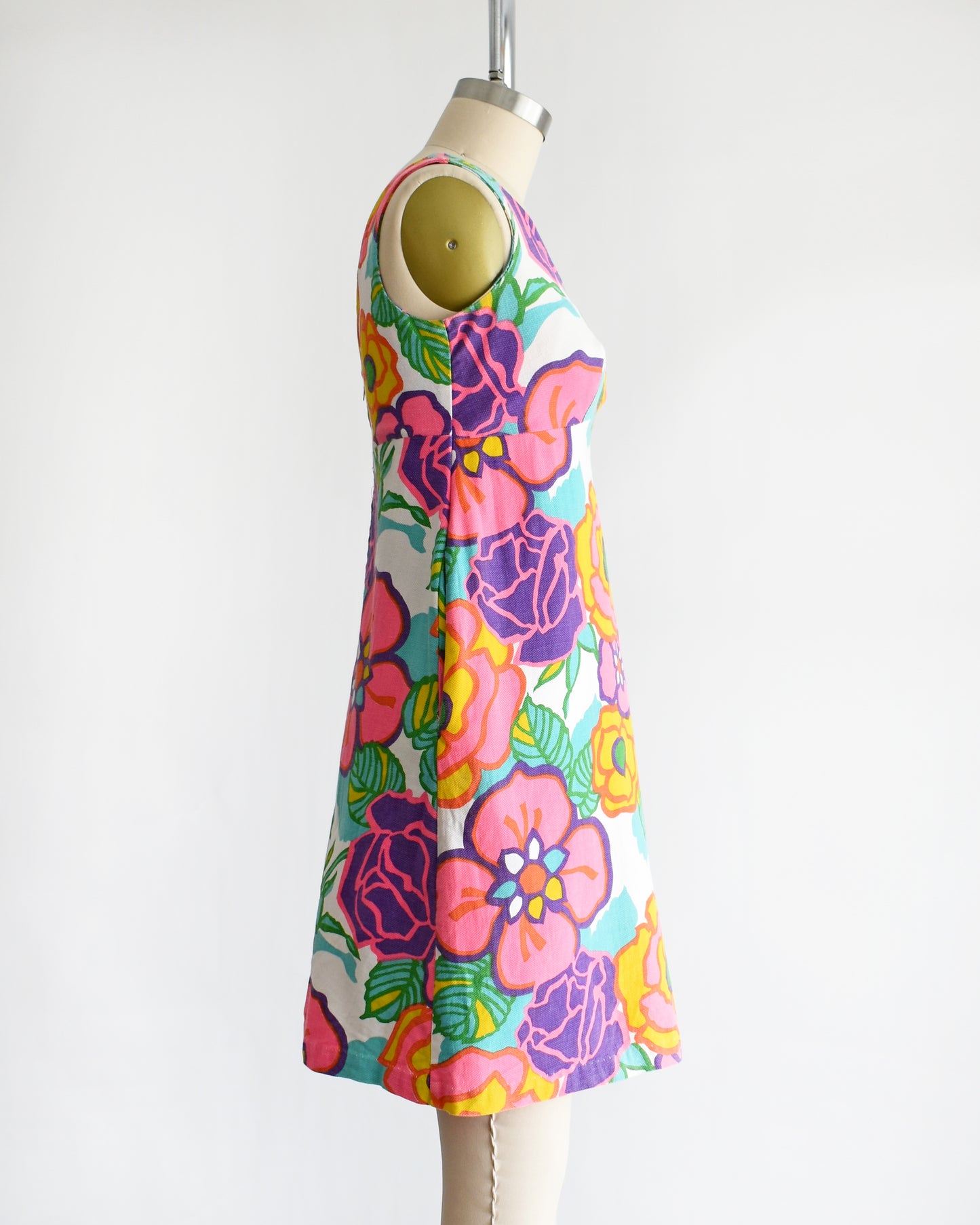 side view of a vintage 1960s sleeveless A-line dress with a vibrant retro floral print, displayed on a cream-colored dress form mannequin against a plain white background.