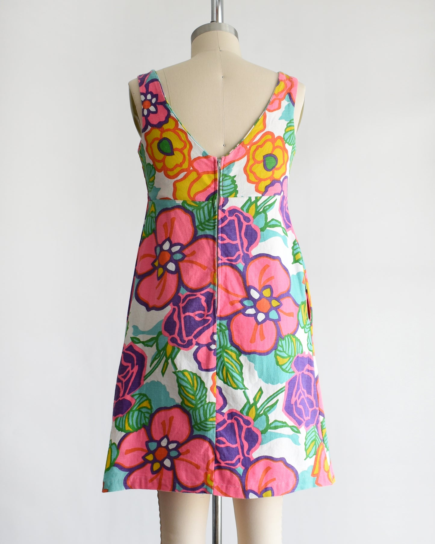 back view of a vintage 1960s sleeveless A-line dress with a vibrant retro floral print, displayed on a cream-colored dress form mannequin against a plain white background.