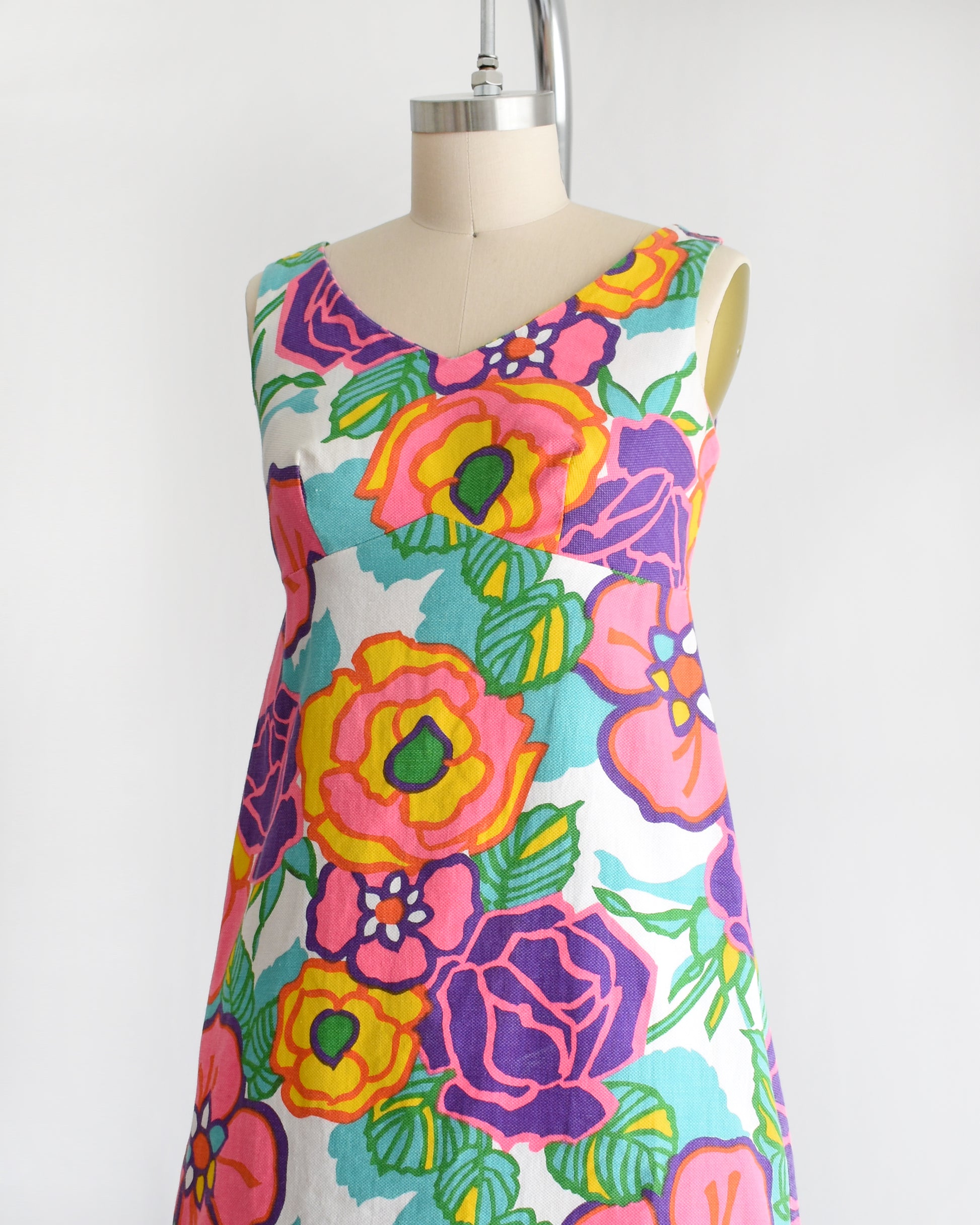 side front view of a vintage 1960s sleeveless A-line dress with a vibrant retro floral print, displayed on a cream-colored dress form mannequin against a plain white background.