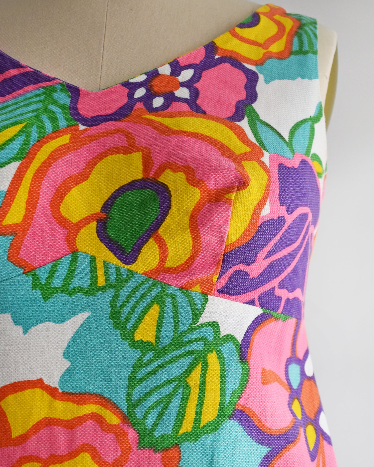 close up of a vintage 1960s sleeveless A-line dress with a vibrant retro floral print, displayed on a cream-colored dress form mannequin against a plain white background.