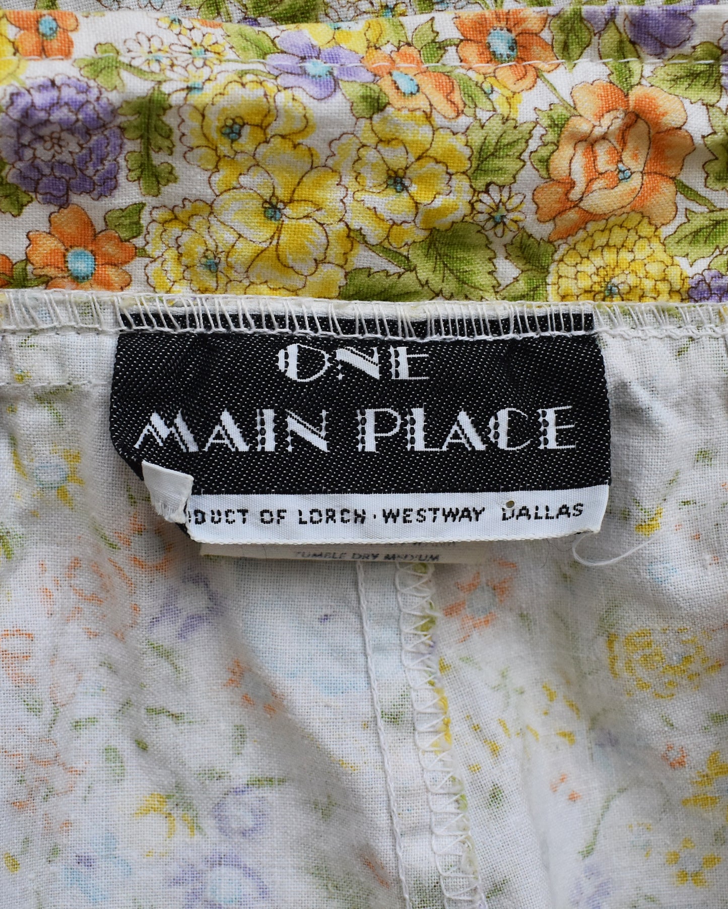 Close-up of the garment label reading ONE MAIN PLACE, with a black brand tag sewn onto white fabric with yellow, orange, and purple floral print.