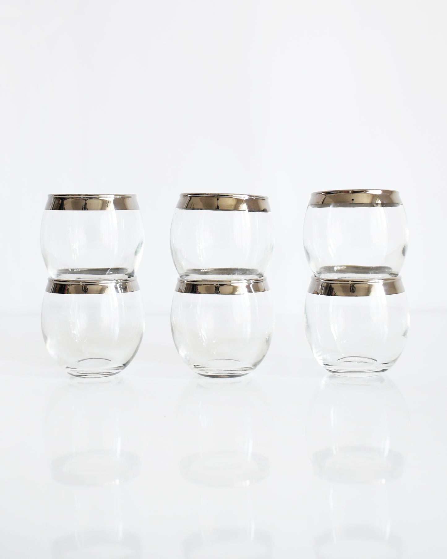 Six clear roly poly glasses with metallic silver bands near the rims, arranged in two rows of three on a reflective white surface against a white background.