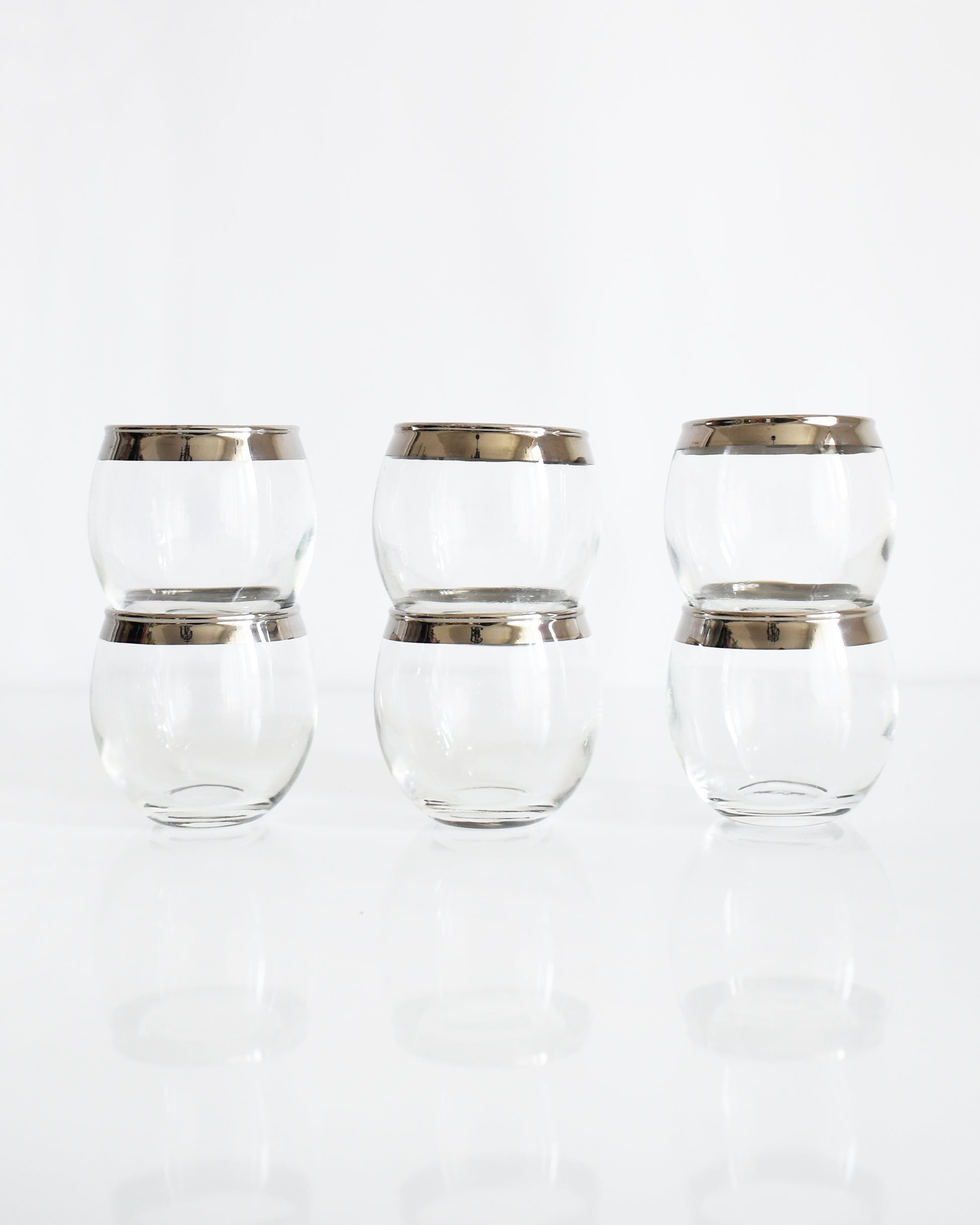Six clear roly poly glasses with metallic silver bands near the rims, arranged in two rows of three on a reflective white surface against a white background.