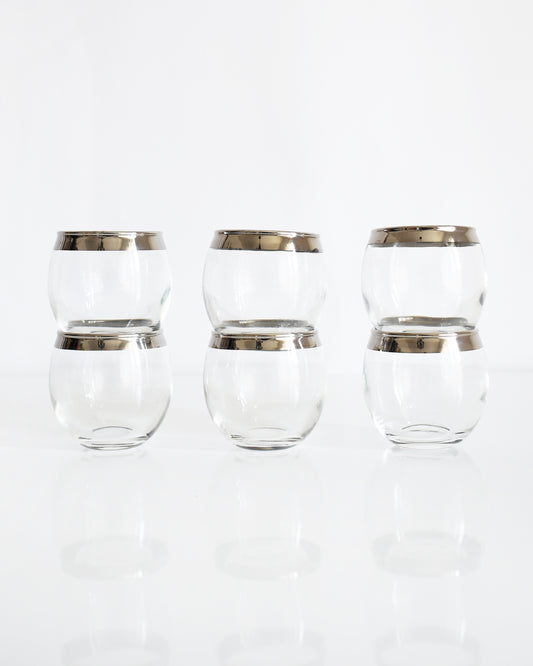 Six clear roly poly glasses with metallic silver bands near the rims, arranged in two rows of three on a reflective white surface against a white background.