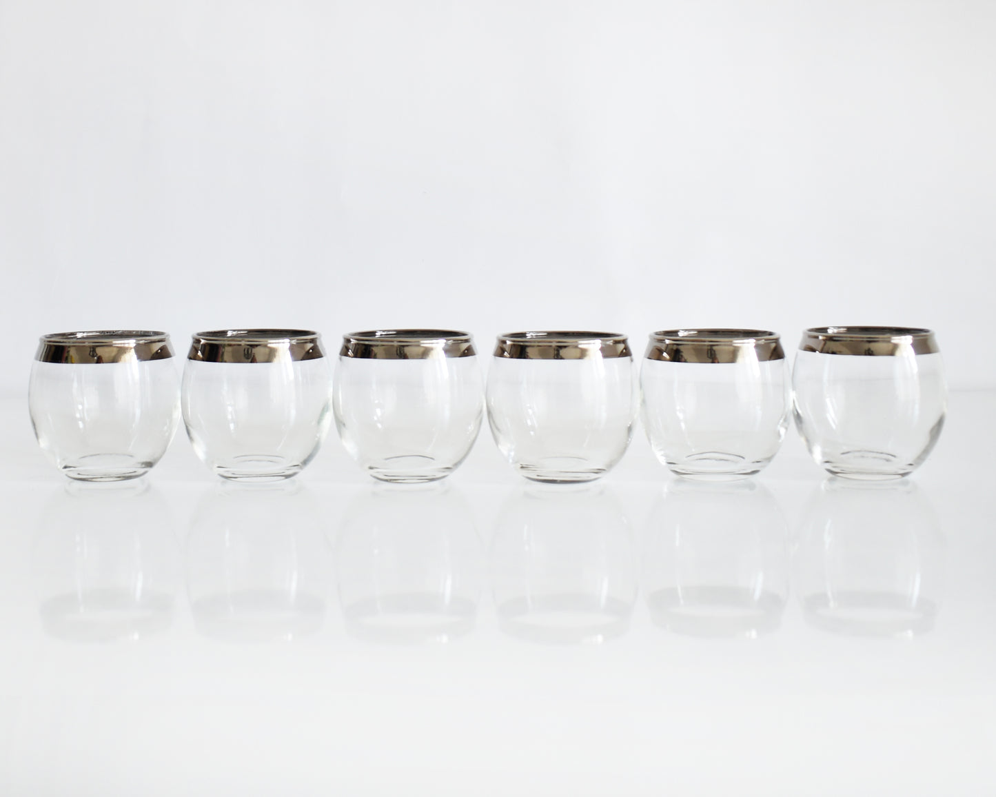 Six clear roly poly glasses with metallic silver bands near the rims, arranged in a row on a reflective white surface against a white background.