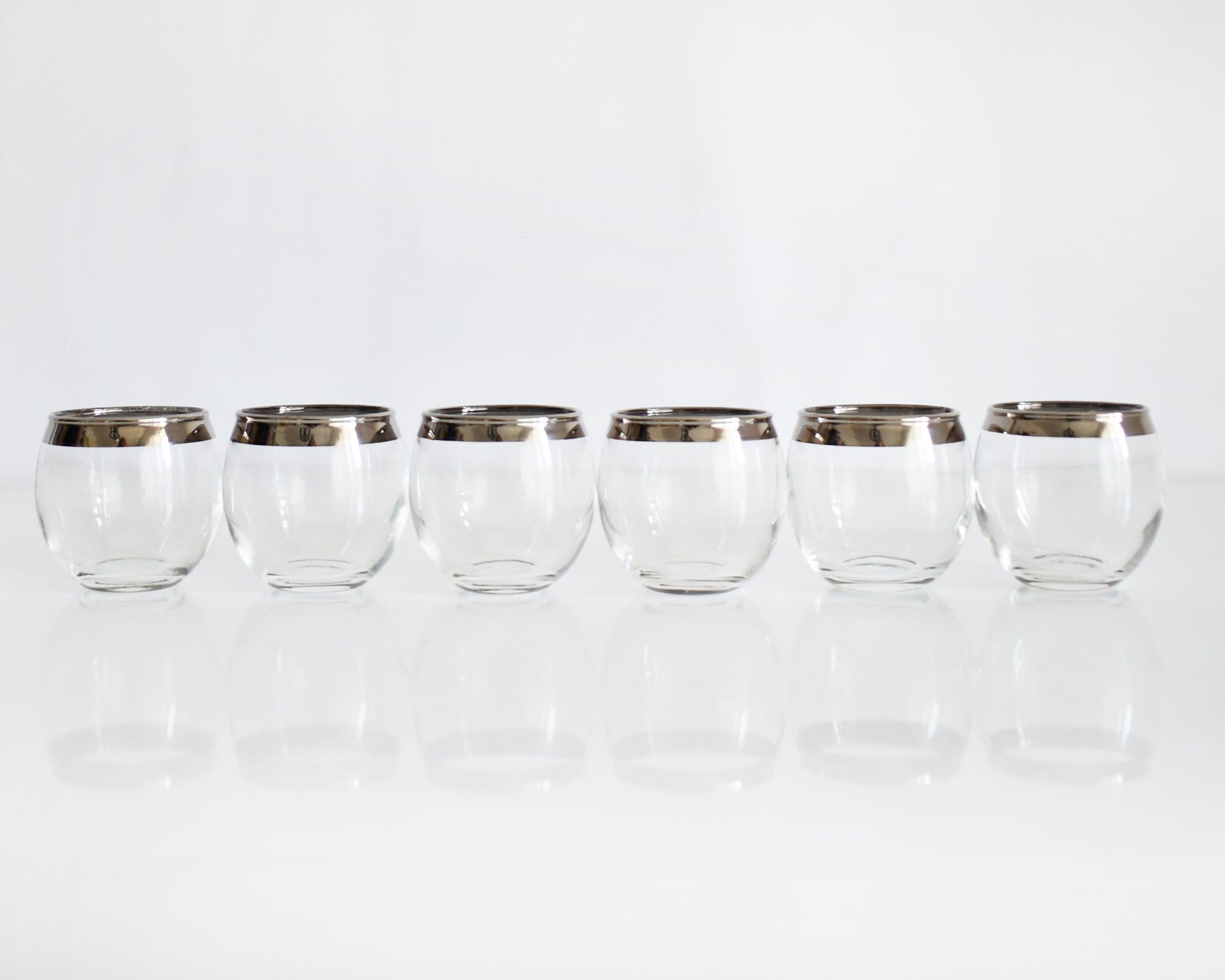 Six clear roly poly glasses with metallic silver bands near the rims, arranged in a row on a reflective white surface against a white background.