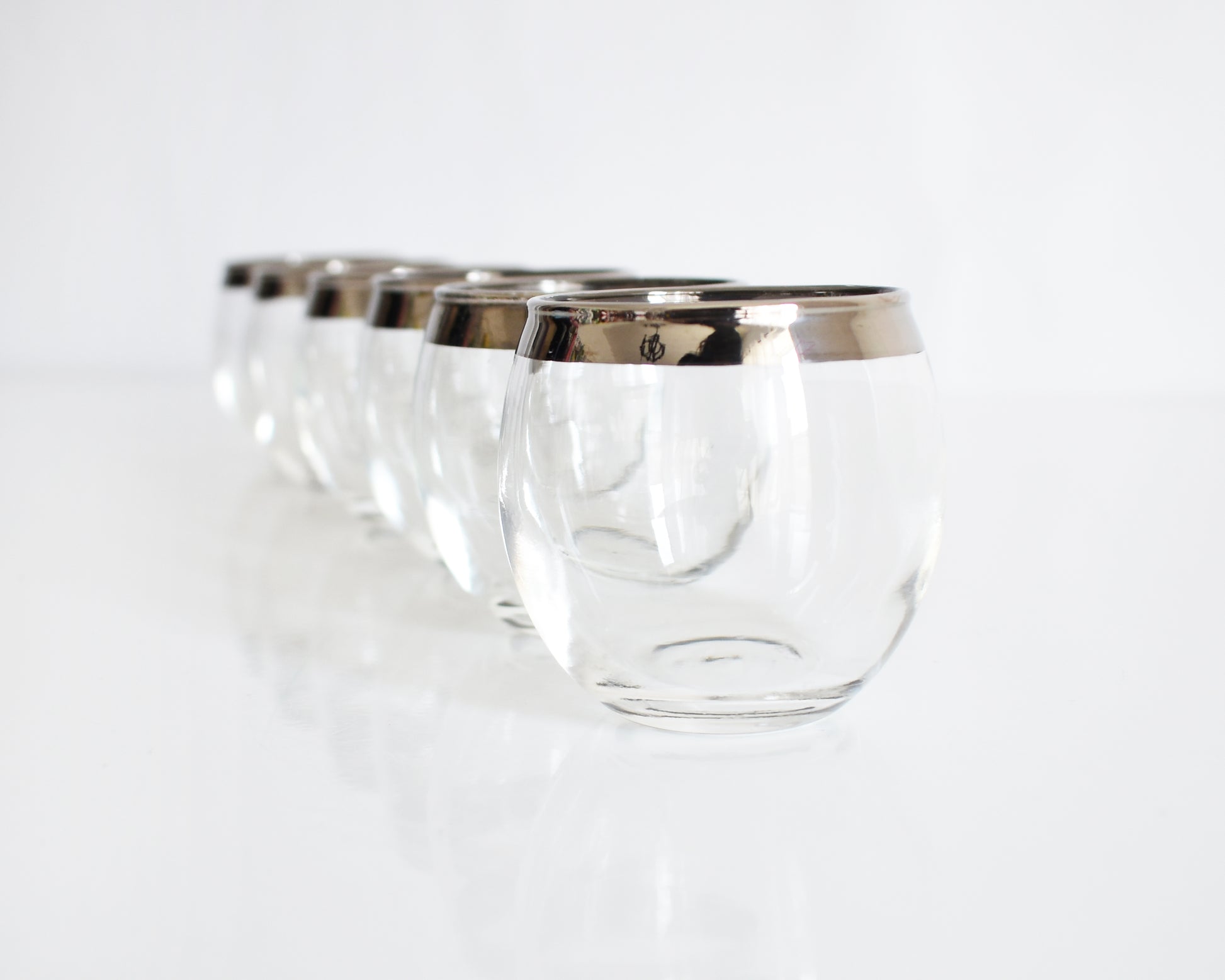 A set of six clear roly poly shot glasses with polished silver bands around the rims, arranged in a diagonal line on a reflective white surface.