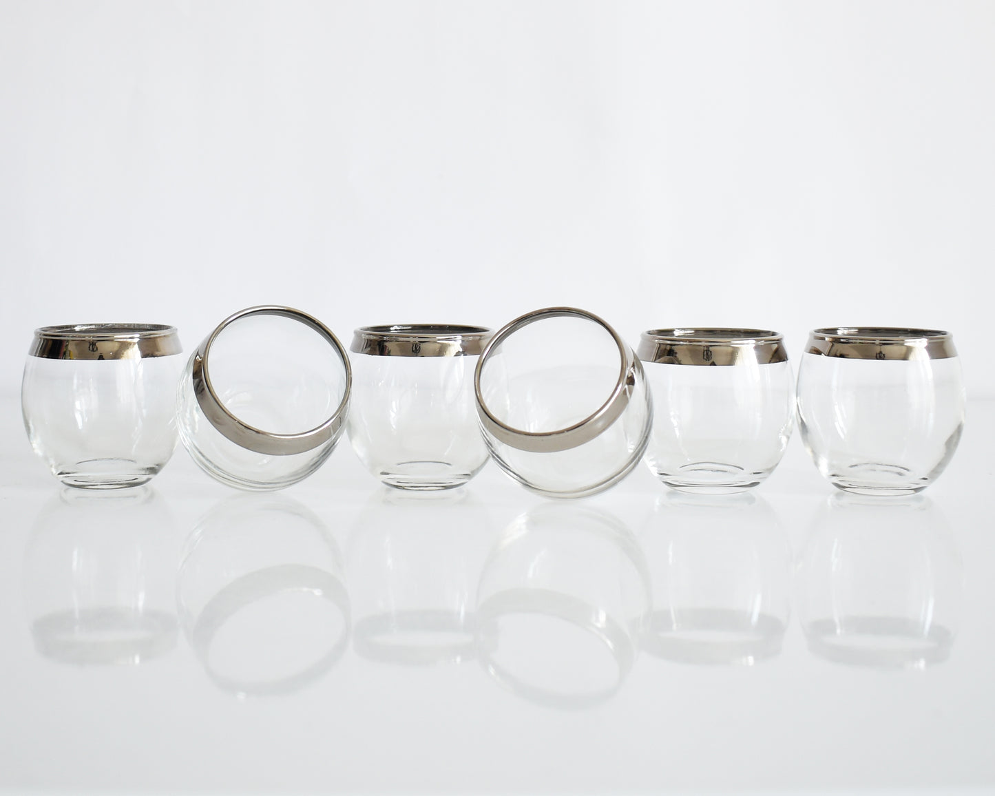 Six clear roly poly glasses with silver metal rims, arranged in a row on a reflective white surface against a white background.