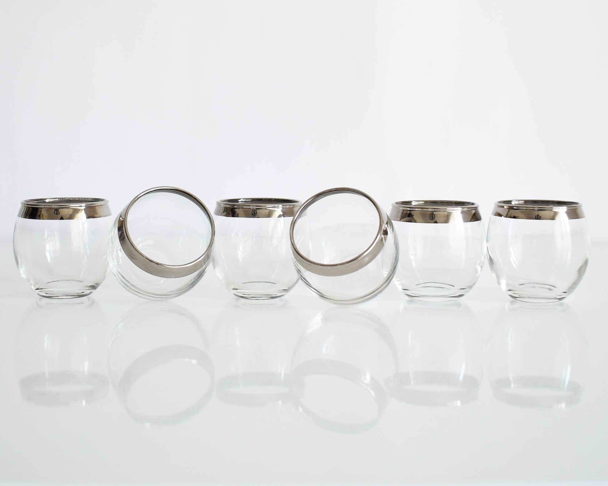 Six clear roly poly glasses with silver metal rims, arranged in a row on a reflective white surface against a white background.