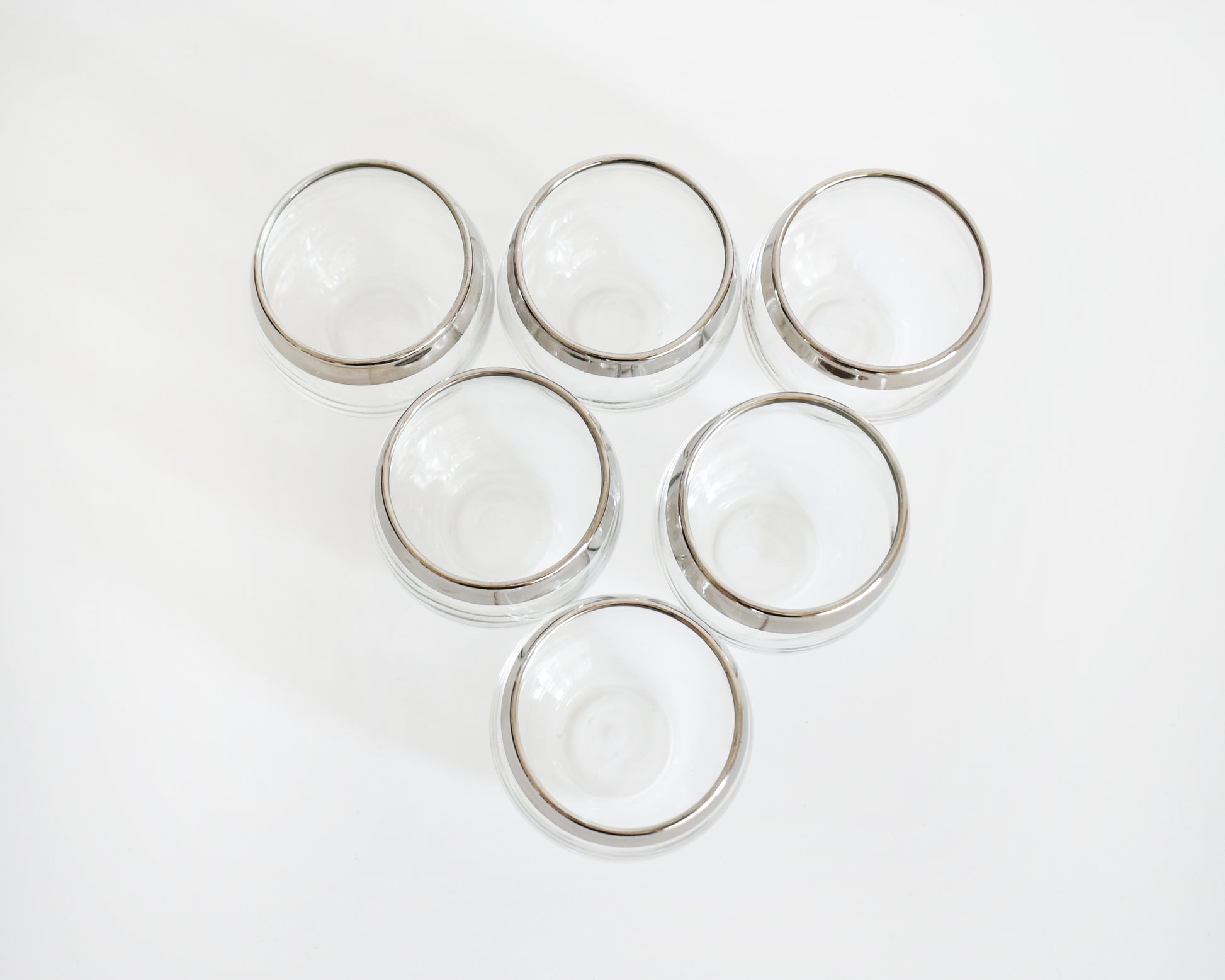 overhead shot of six clear roly poly glasses with polished silver rims, arranged in rows on a plain white background.