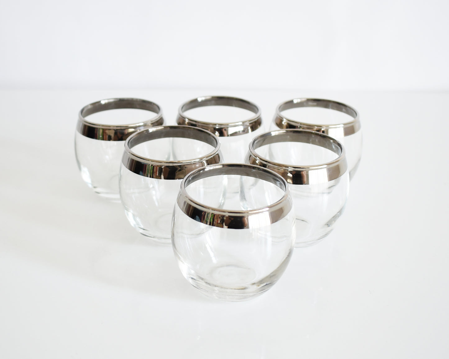 Six clear roly poly glass cocktail tumblers with silver metal rims, arranged on a clean white surface against a white background.