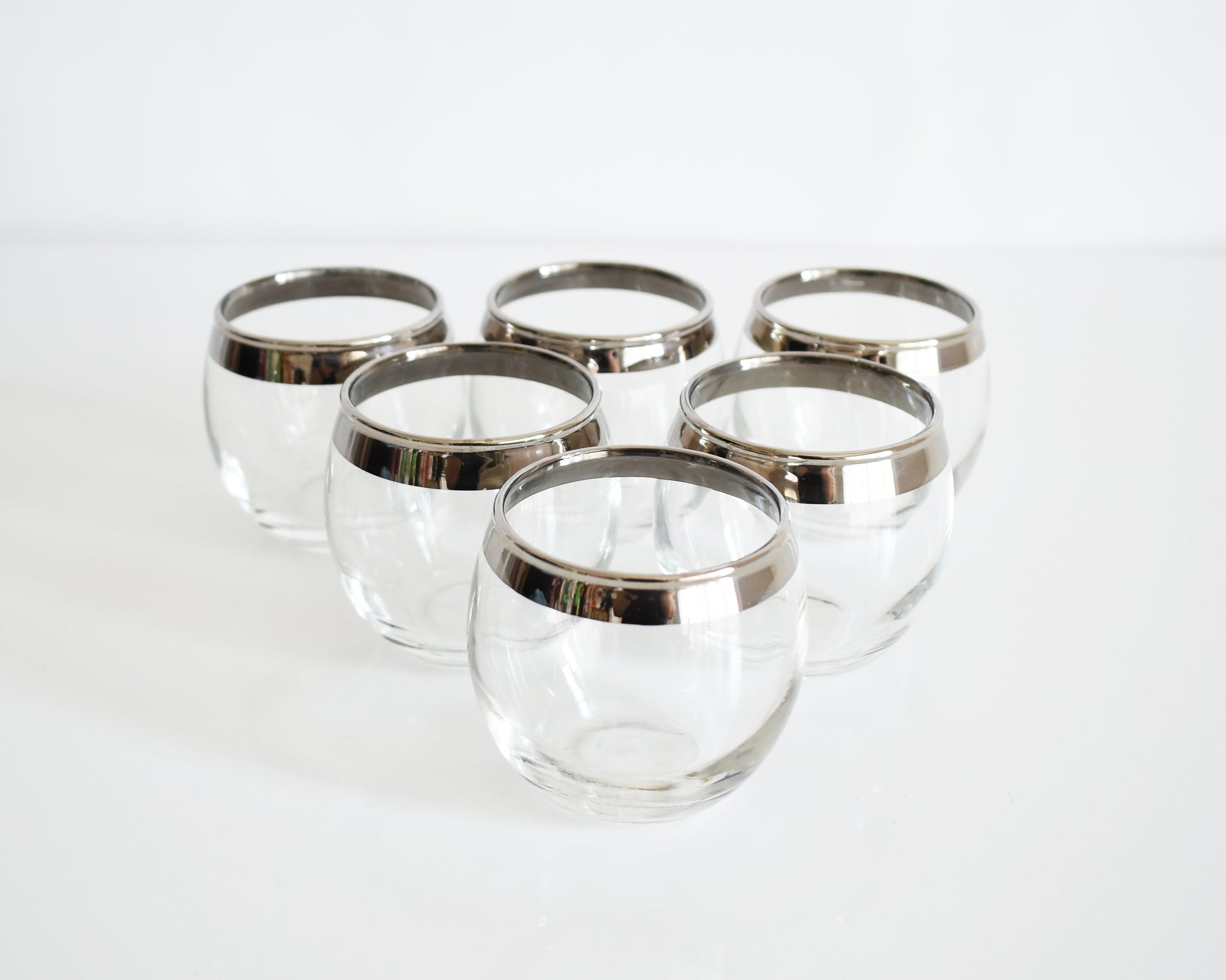 Six clear roly poly glass cocktail tumblers with silver metal rims, arranged on a clean white surface against a white background.