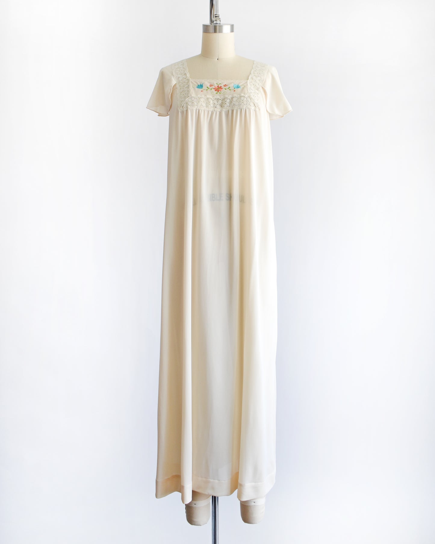 a vintage 1970s cream-colored nightgown with short sleeves and delicate floral embroidery across the chest, displayed on a dress form mannequin against a white background.