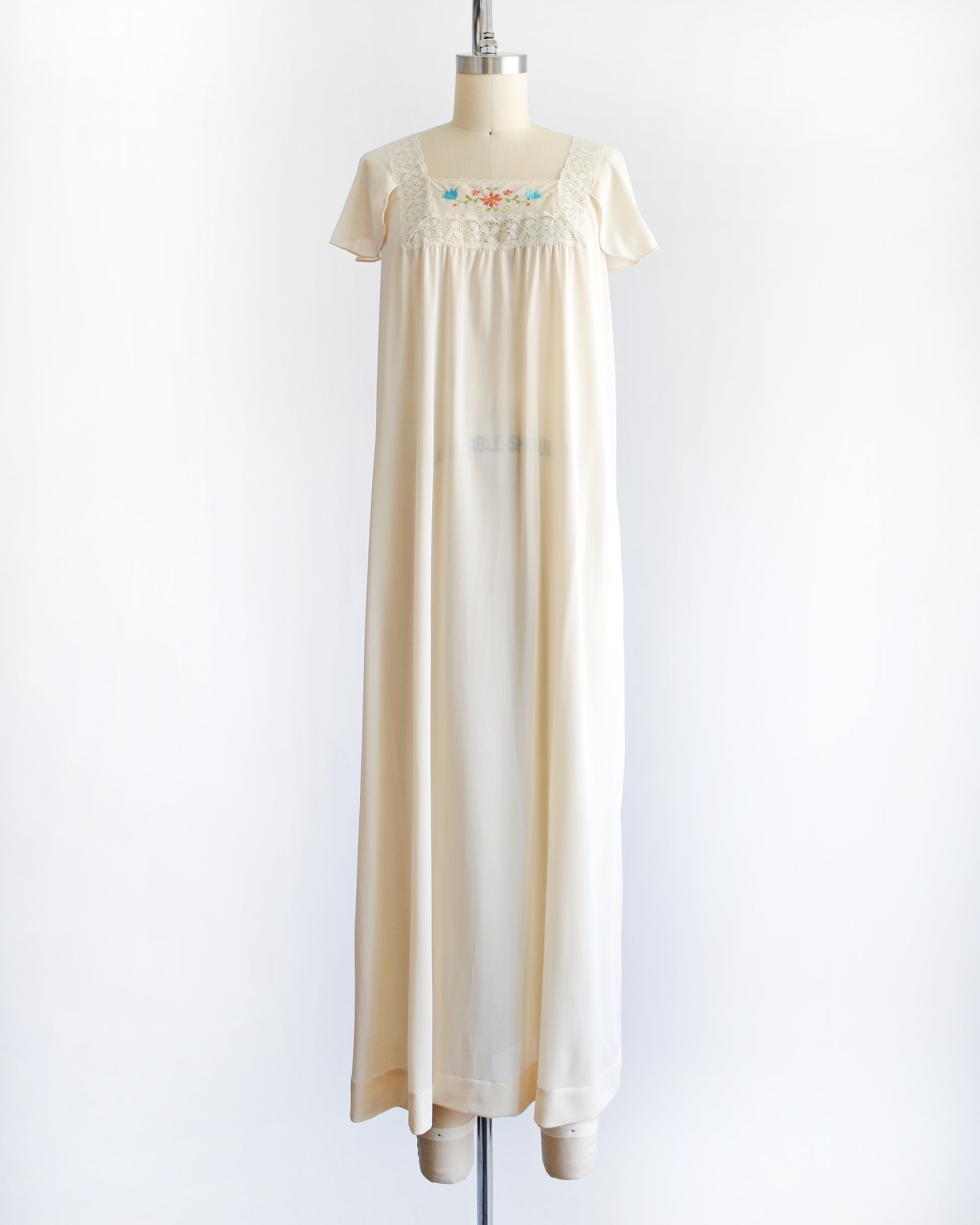a vintage 1970s cream-colored nightgown with short sleeves and delicate floral embroidery across the chest, displayed on a dress form mannequin against a white background.