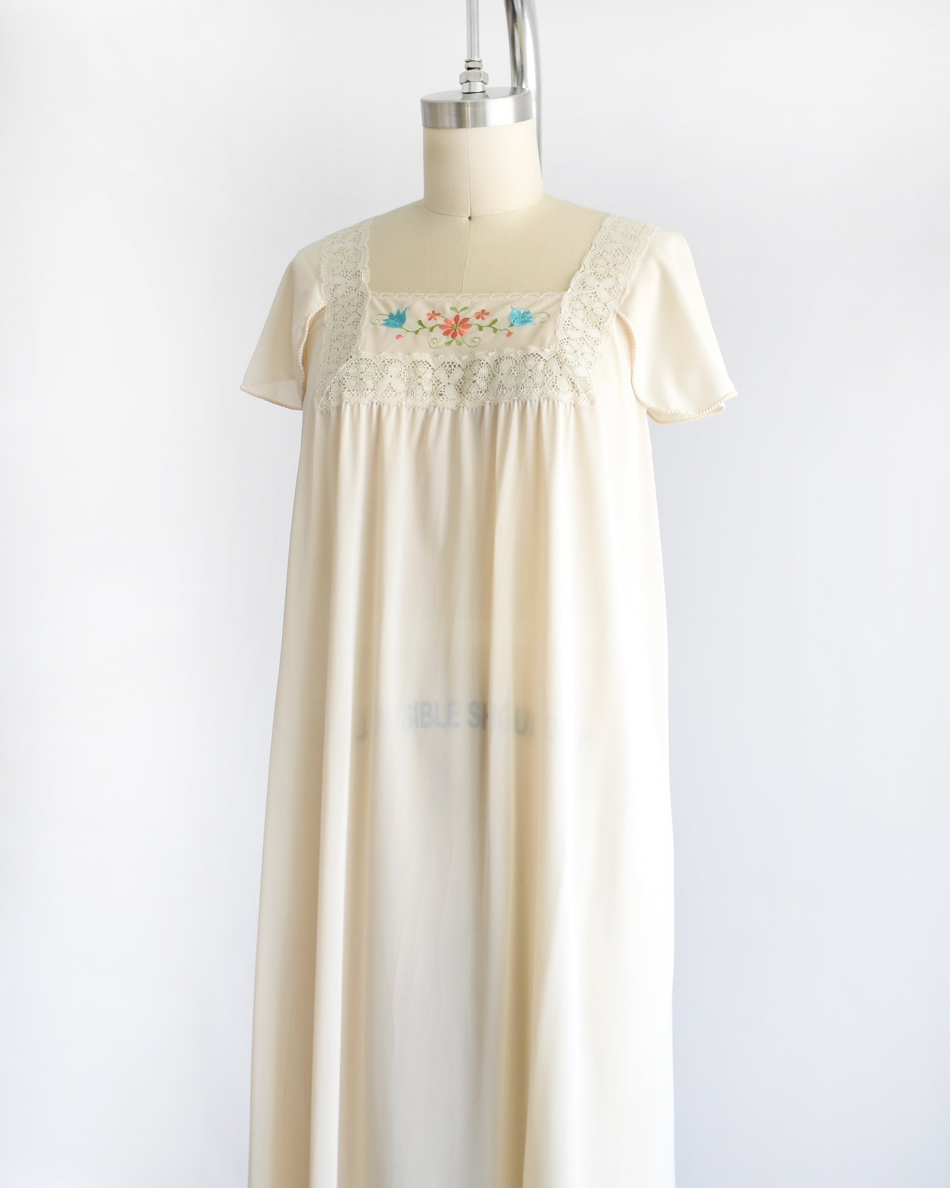 side front view of a vintage 1970s cream-colored nightgown with short sleeves and delicate floral embroidery across the chest, displayed on a dress form mannequin against a white background.