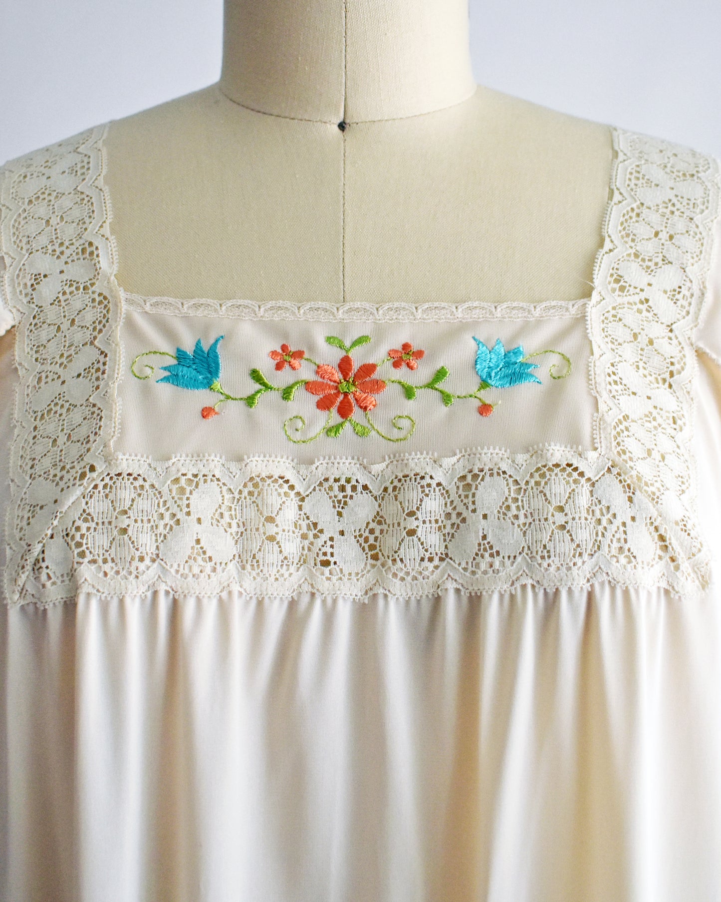 close up of the lace and embroidered detail on a vintage 1970s cream-colored nightgown with short sleeves and delicate floral embroidery across the chest, displayed on a dress form mannequin against a white background.