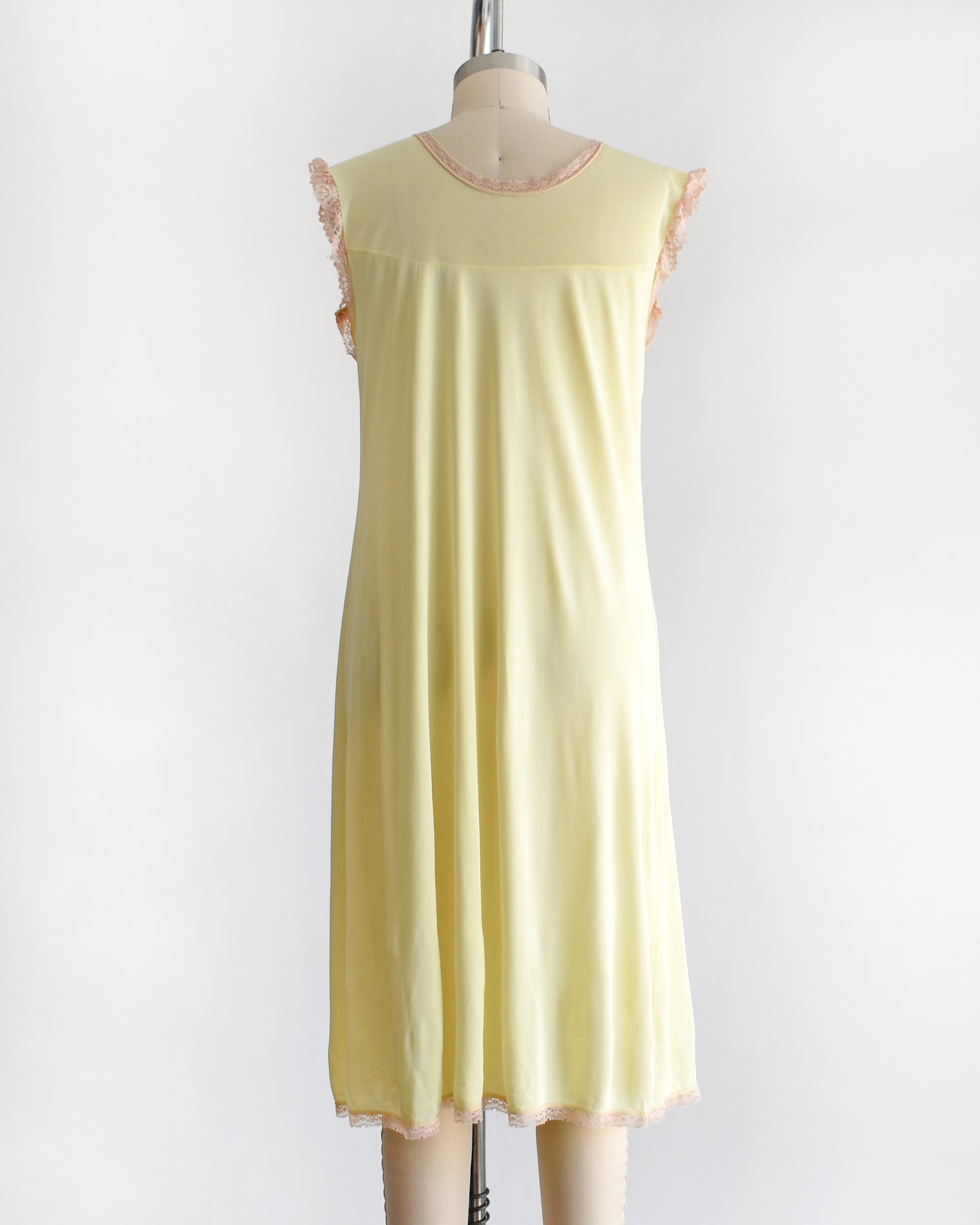 back view of a vintage 1960s pale yellow slip-style nightgown with a peach lace yoke and ruffled trim, displayed on a mannequin against a plain white background.