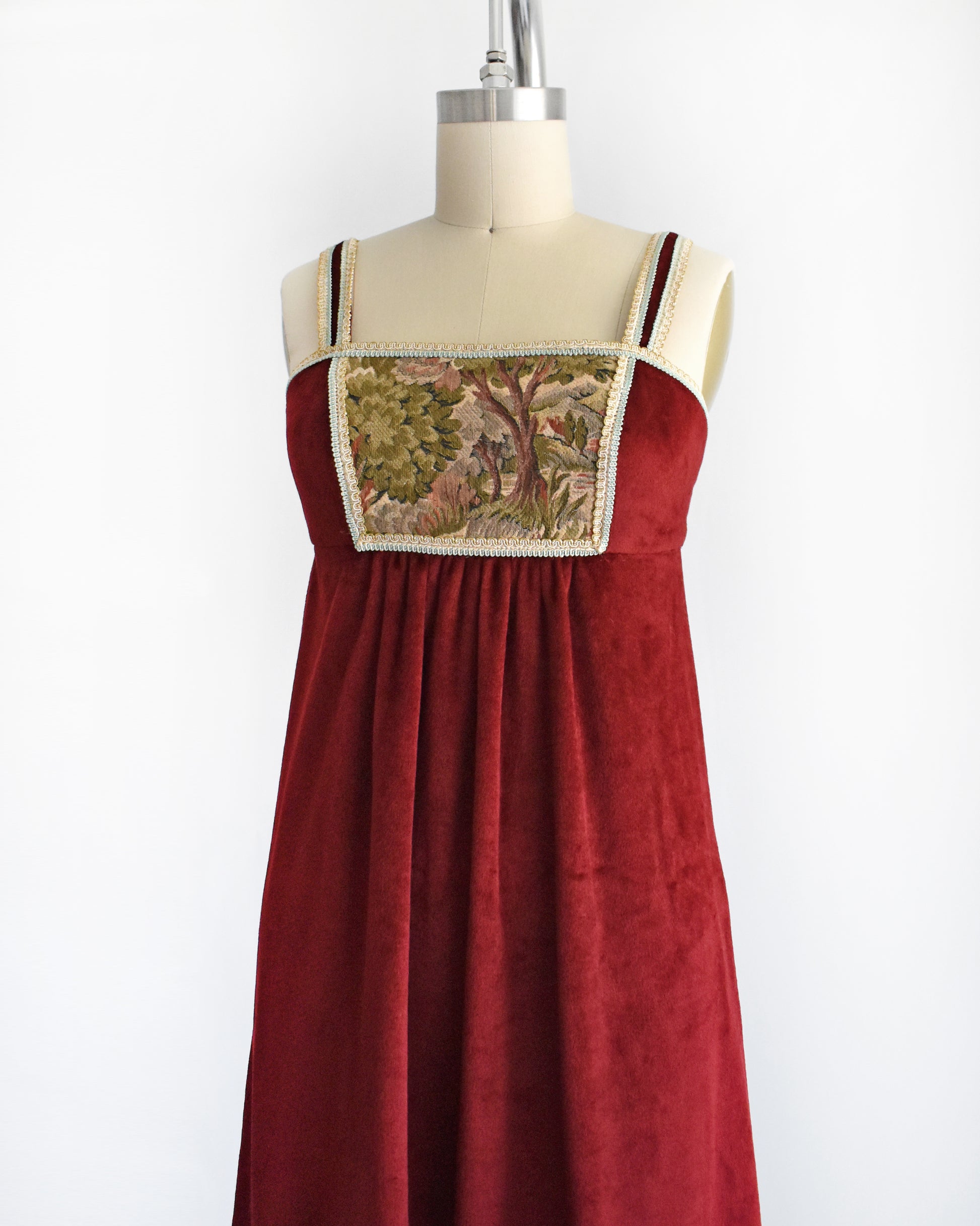 side front view of A vintage 1970s burgundy velvet maxi dress that has a woodsy tapestry scene on the bodice.