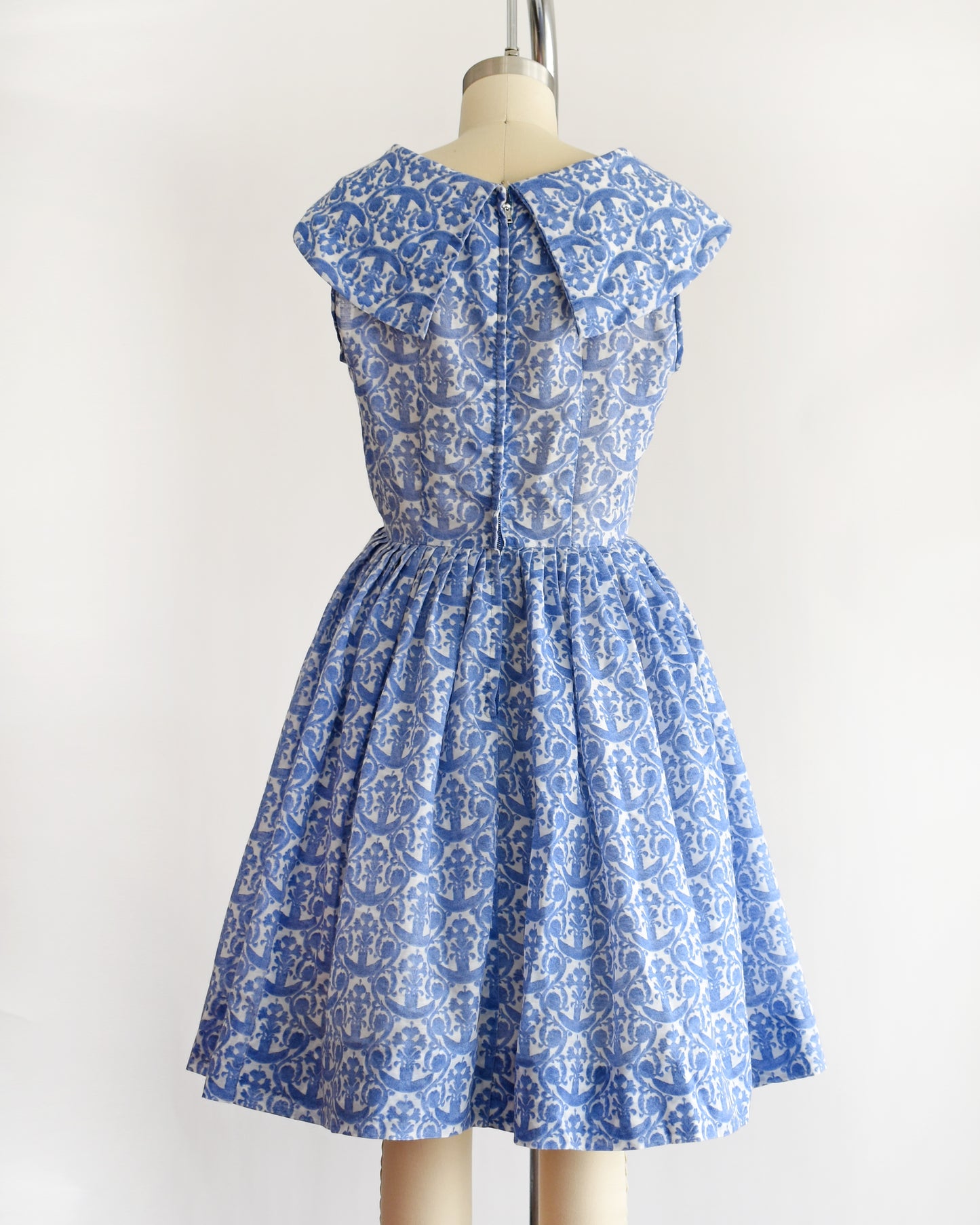 back view of a vintage 1960s blue and white patterned dress displayed on a beige dress form mannequin against a plain white background.