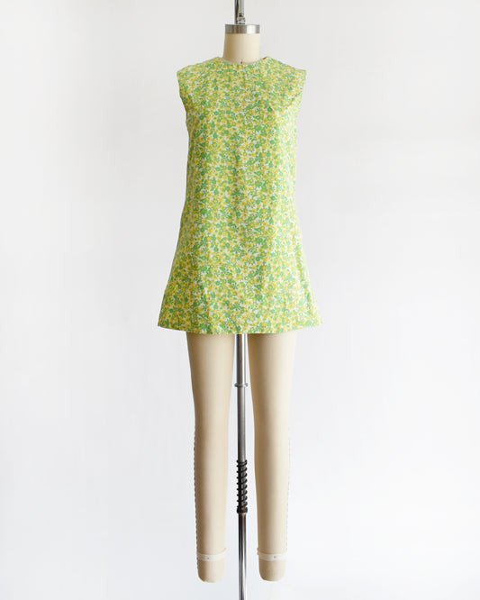 A  vintage 1960s sleeveless, A-line mini dress with a green and yellow floral pattern, displayed on a beige mannequin against a plain white background.