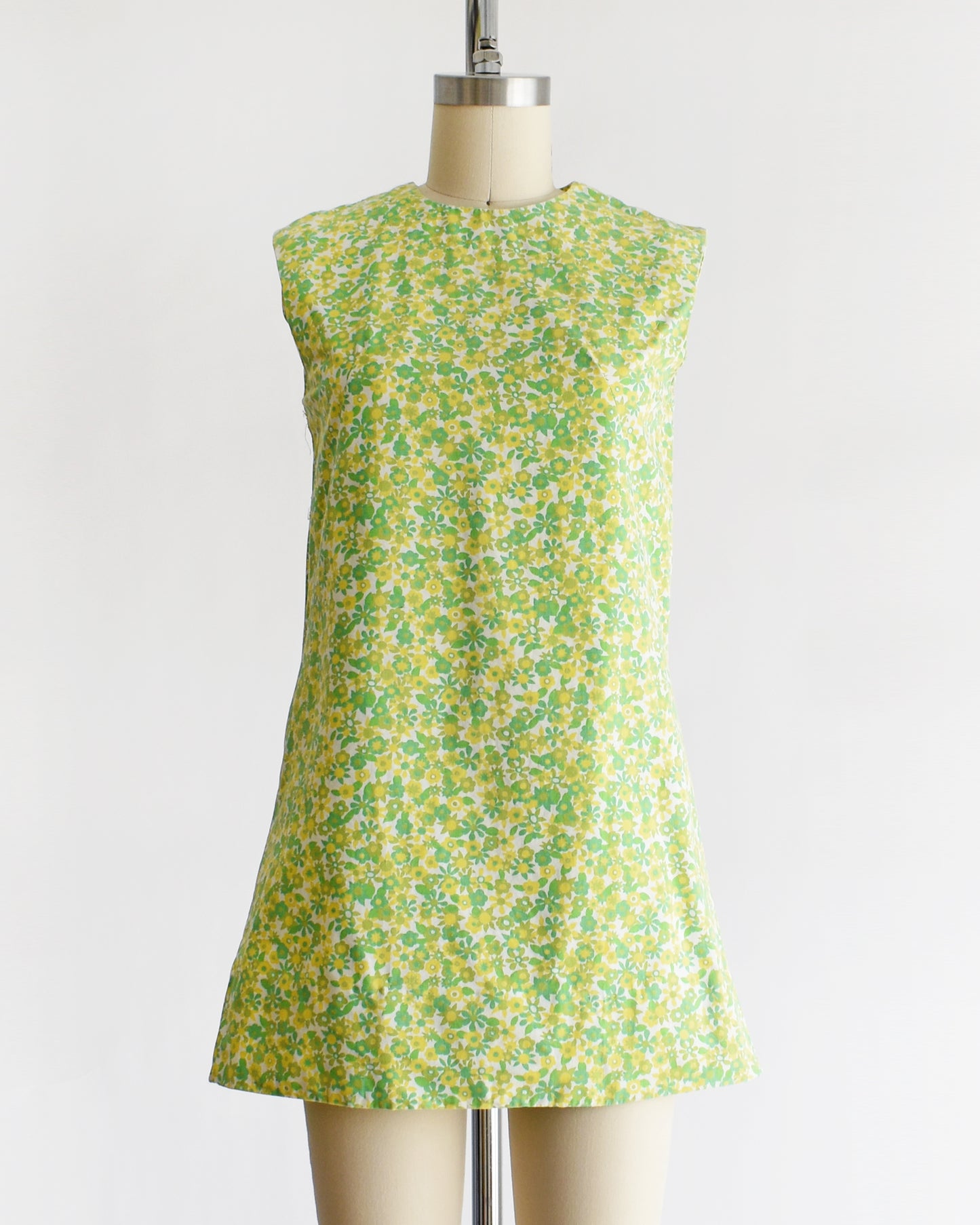 A  vintage 1960s sleeveless, A-line mini dress with a green and yellow floral pattern, displayed on a beige mannequin against a plain white background.