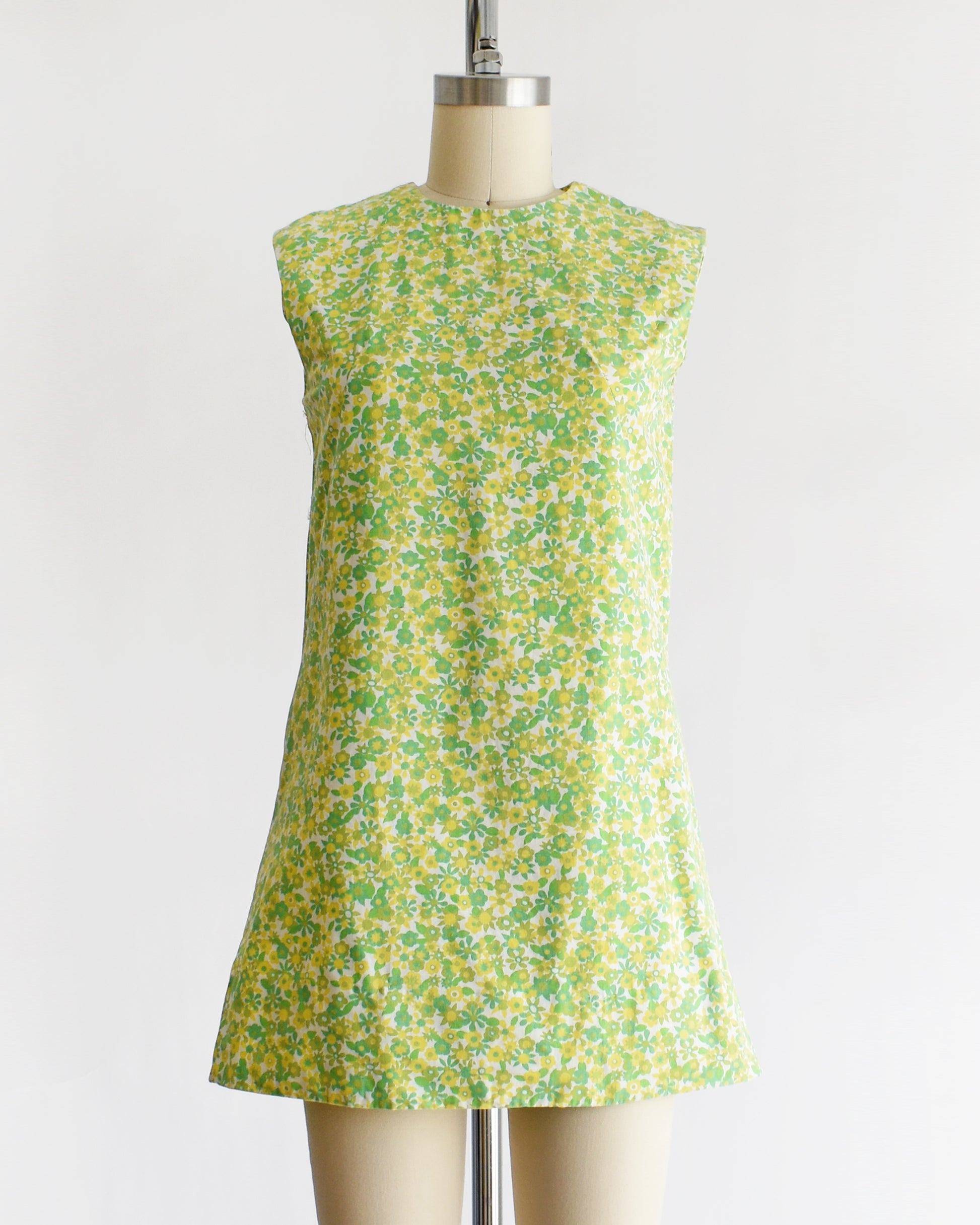 A  vintage 1960s sleeveless, A-line mini dress with a green and yellow floral pattern, displayed on a beige mannequin against a plain white background.