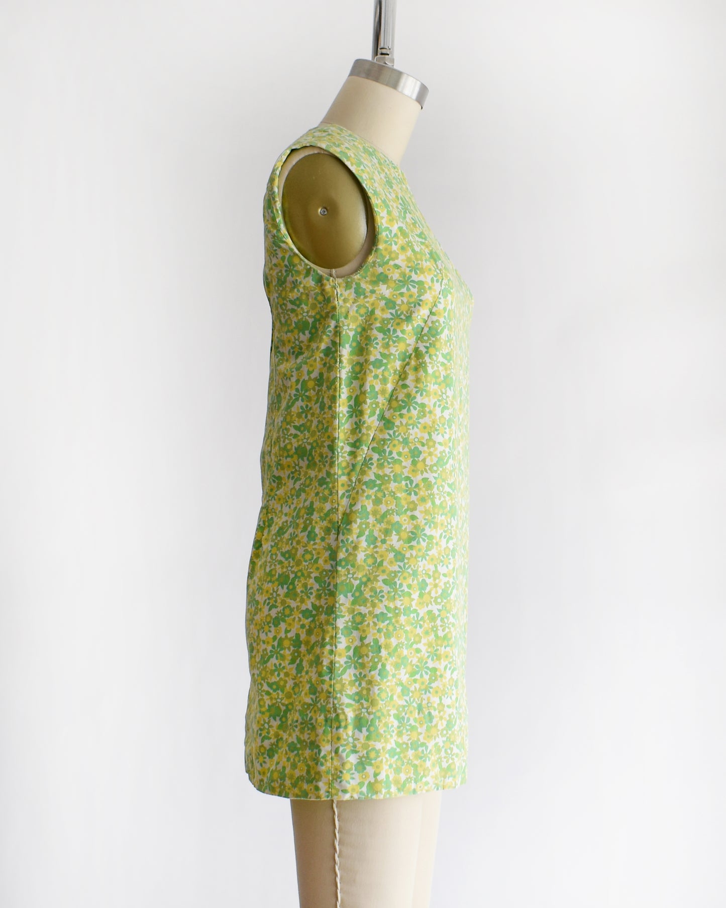 side view of a  vintage 1960s sleeveless, A-line mini dress with a green and yellow floral pattern, displayed on a beige mannequin against a plain white background.
