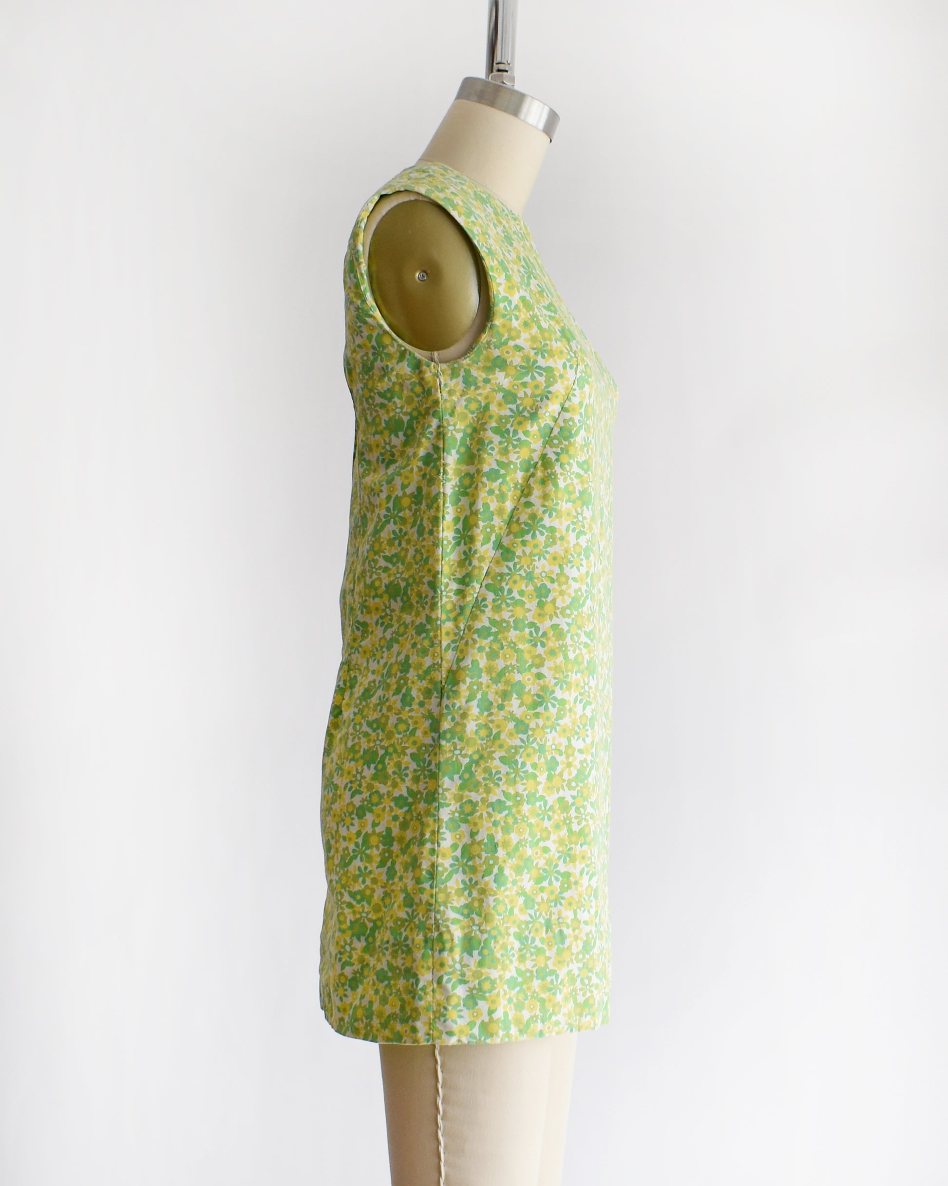 side view of a  vintage 1960s sleeveless, A-line mini dress with a green and yellow floral pattern, displayed on a beige mannequin against a plain white background.