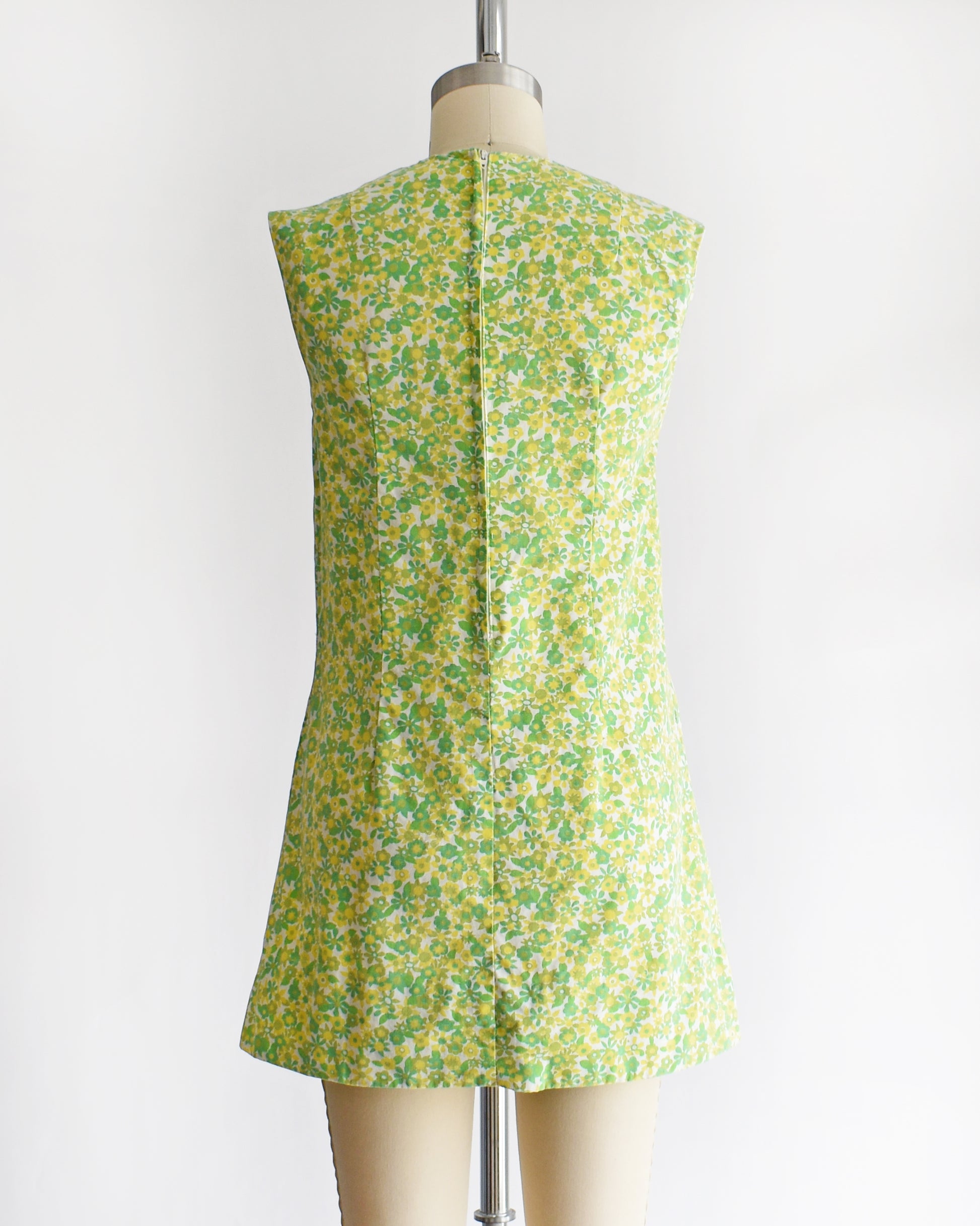 back view of a  vintage 1960s sleeveless, A-line mini dress with a green and yellow floral pattern, displayed on a beige mannequin against a plain white background.