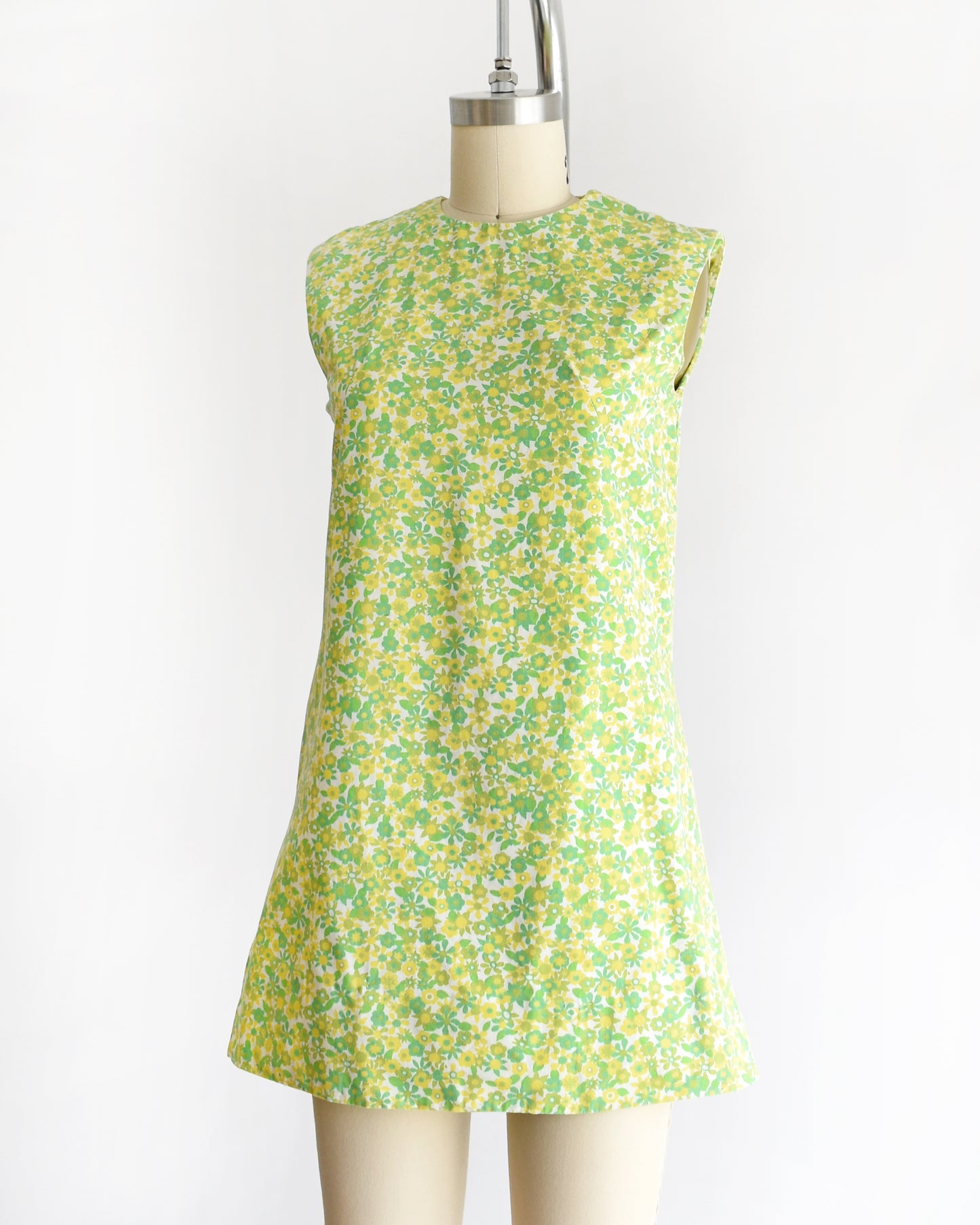 side front view of a  vintage 1960s sleeveless, A-line mini dress with a green and yellow floral pattern, displayed on a beige mannequin against a plain white background.