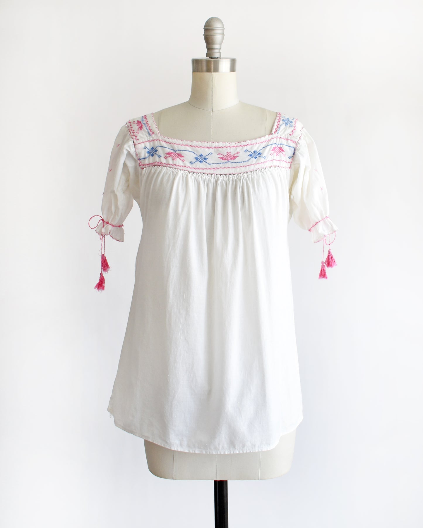 a vintage 1970s white short-sleeve blouse with colorful floral embroidery across the neckline, tied bow details on the sleeves, and pink tassels, displayed on a cream dress form against a white background.