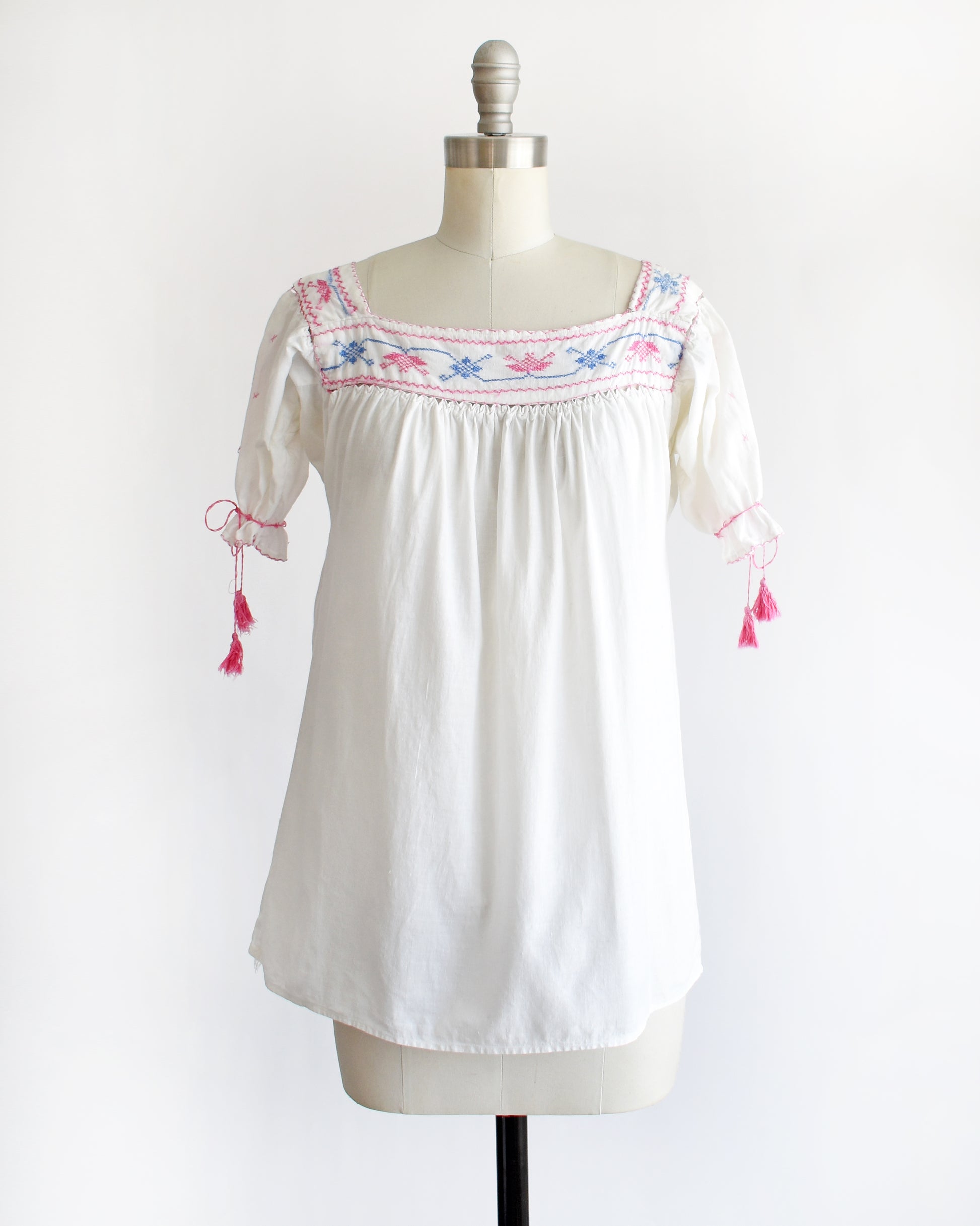 a vintage 1970s white short-sleeve blouse with colorful floral embroidery across the neckline, tied bow details on the sleeves, and pink tassels, displayed on a cream dress form against a white background.
