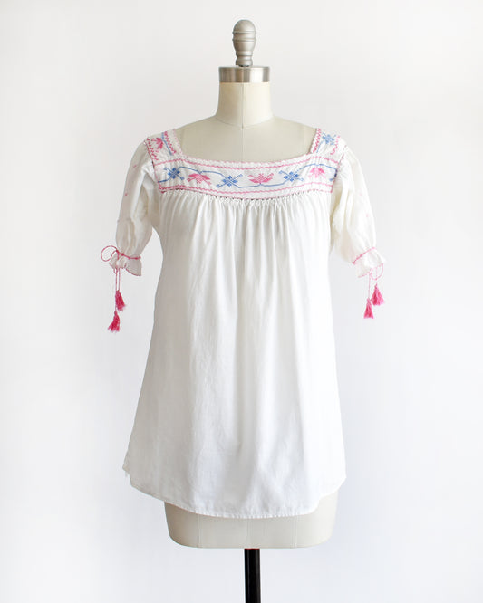 a vintage 1970s white short-sleeve blouse with colorful floral embroidery across the neckline, tied bow details on the sleeves, and pink tassels, displayed on a cream dress form against a white background.