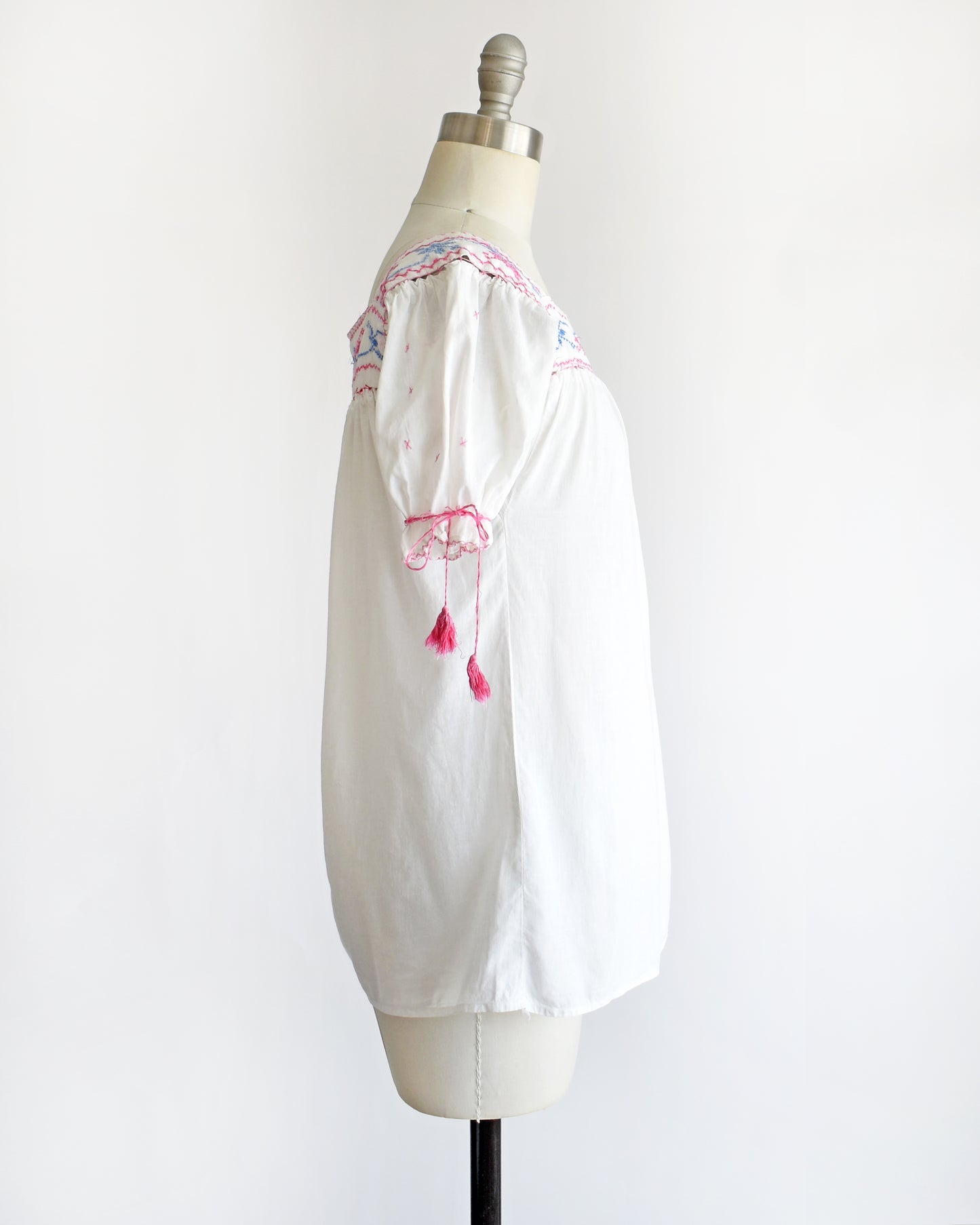 side view of a vintage 1970s white short-sleeve blouse with colorful floral embroidery across the neckline, tied bow details on the sleeves, and pink tassels, displayed on a cream dress form against a white background.