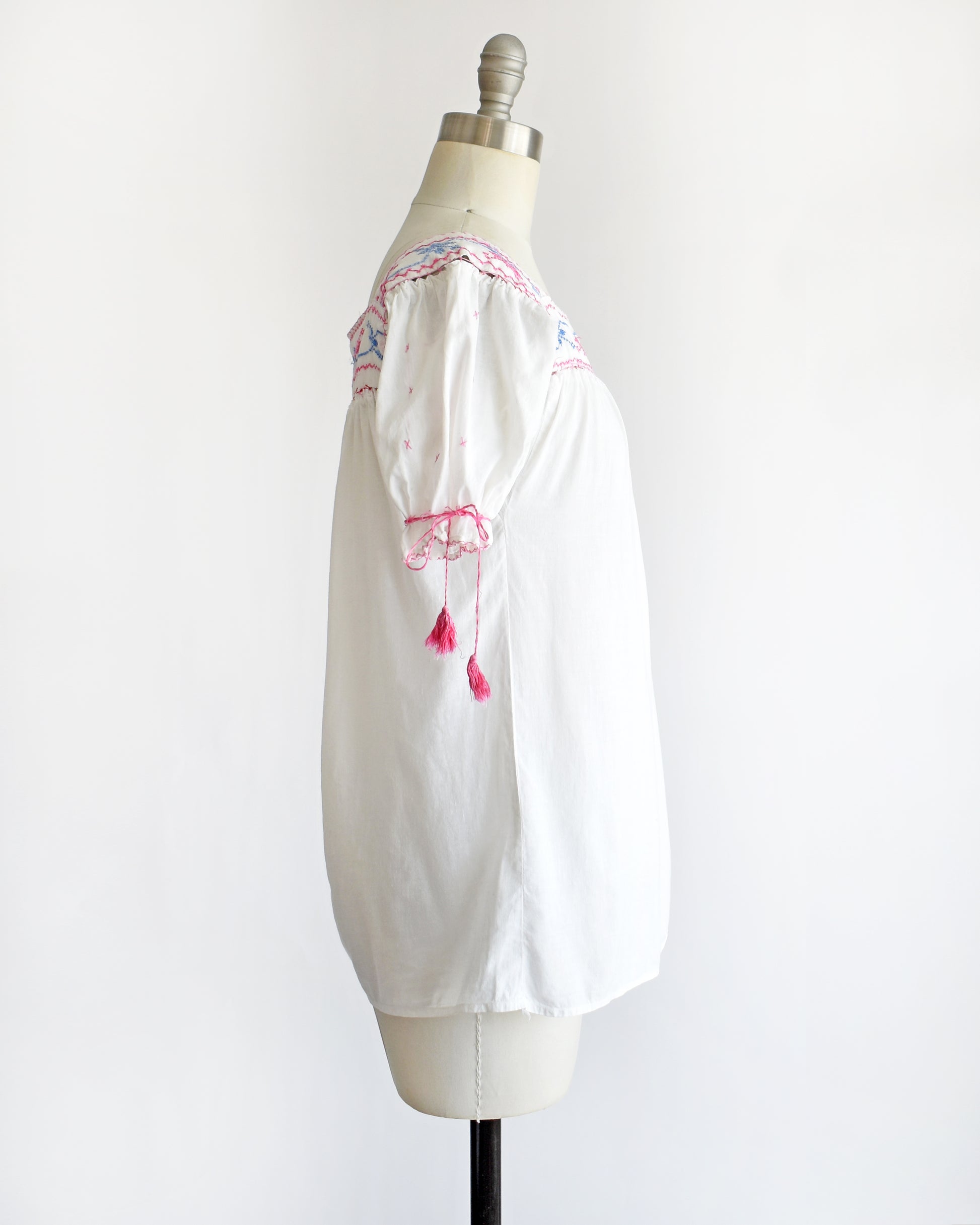 side view of a vintage 1970s white short-sleeve blouse with colorful floral embroidery across the neckline, tied bow details on the sleeves, and pink tassels, displayed on a cream dress form against a white background.
