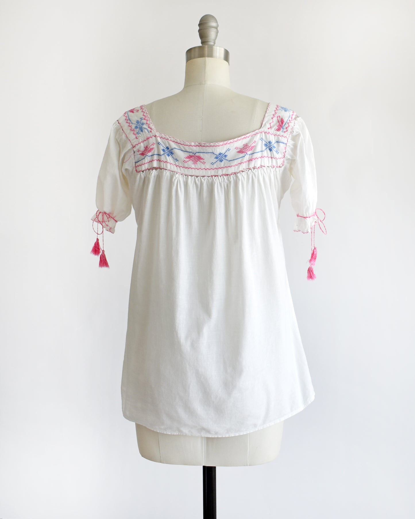 back view of a vintage 1970s white short-sleeve blouse with colorful floral embroidery across the neckline, tied bow details on the sleeves, and pink tassels, displayed on a cream dress form against a white background.