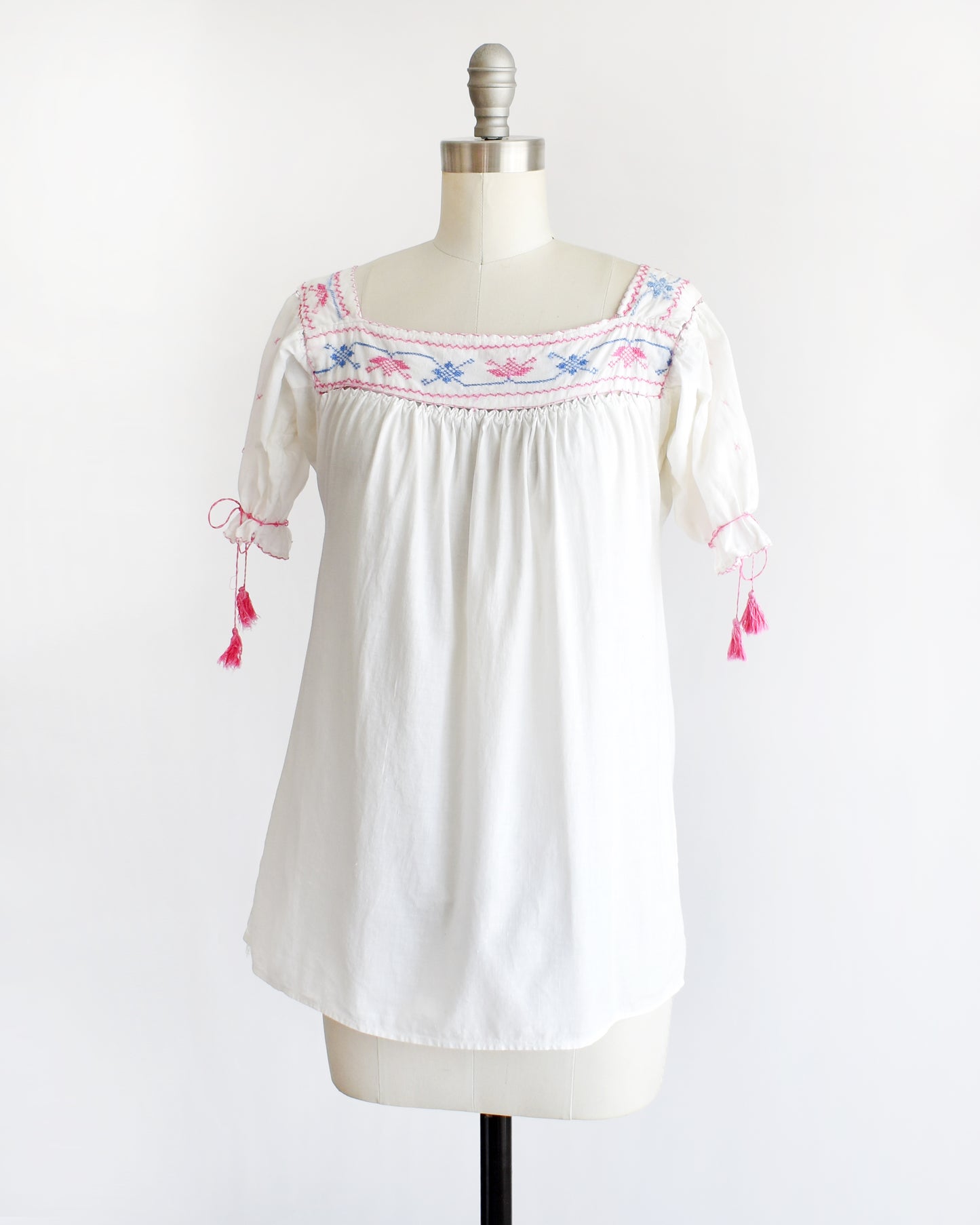 side front view of a vintage 1970s white short-sleeve blouse with colorful floral embroidery across the neckline, tied bow details on the sleeves, and pink tassels, displayed on a cream dress form against a white background.