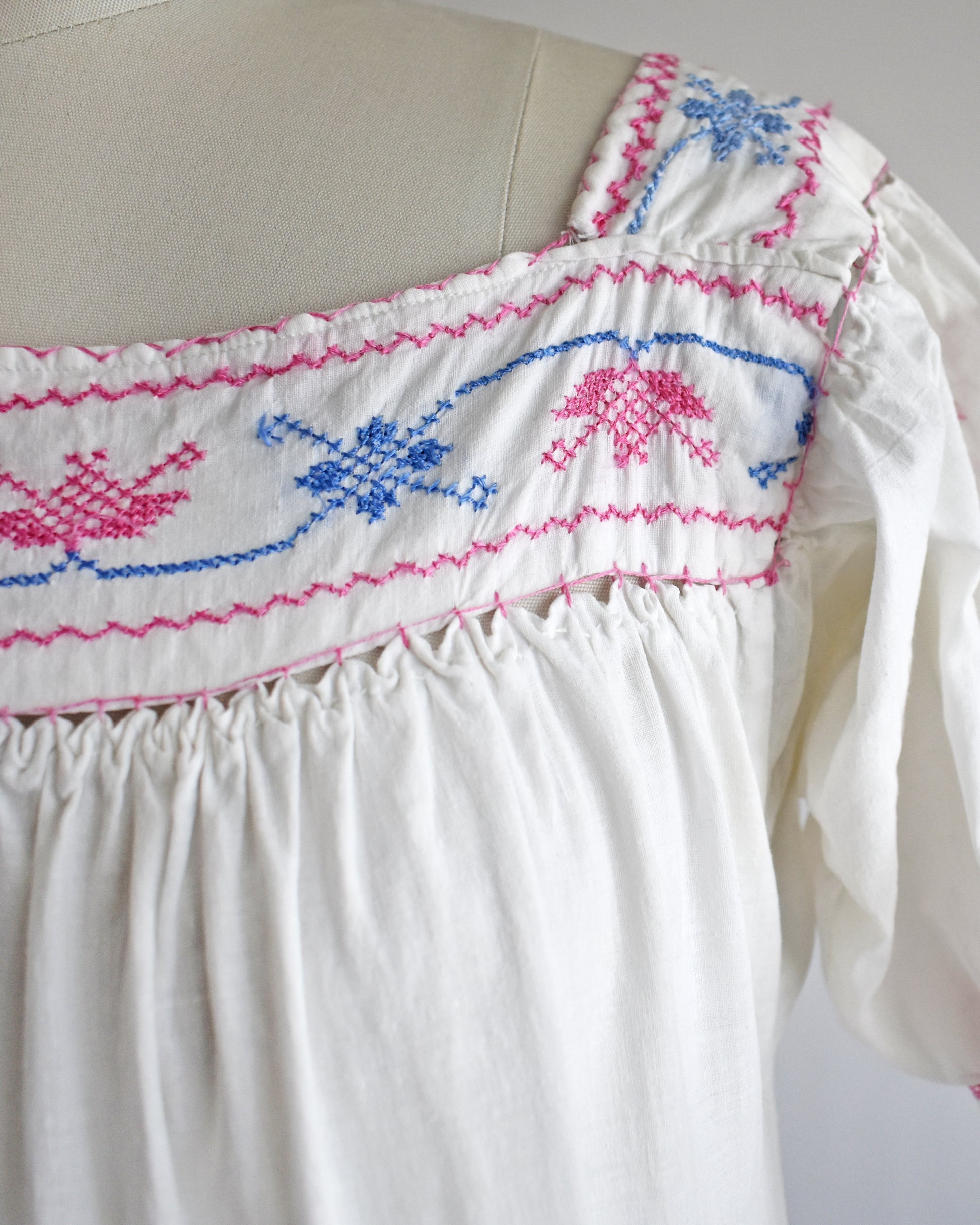 Close-up of a white embroidered blouse on a dress form, featuring pink and blue geometric and floral embroidery across the smocked bodice and shoulder.