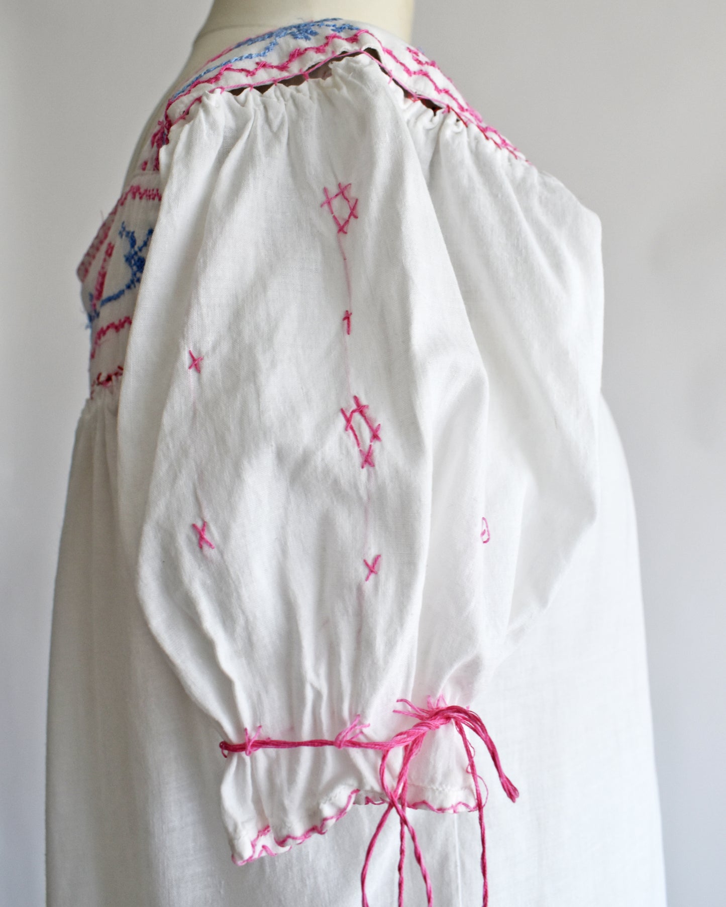 close up of the sleeve on a blouse with pink embroidered details, geometric star patterns, and a pink drawstring tie at the waist, displayed on a mannequin against a plain white background.
