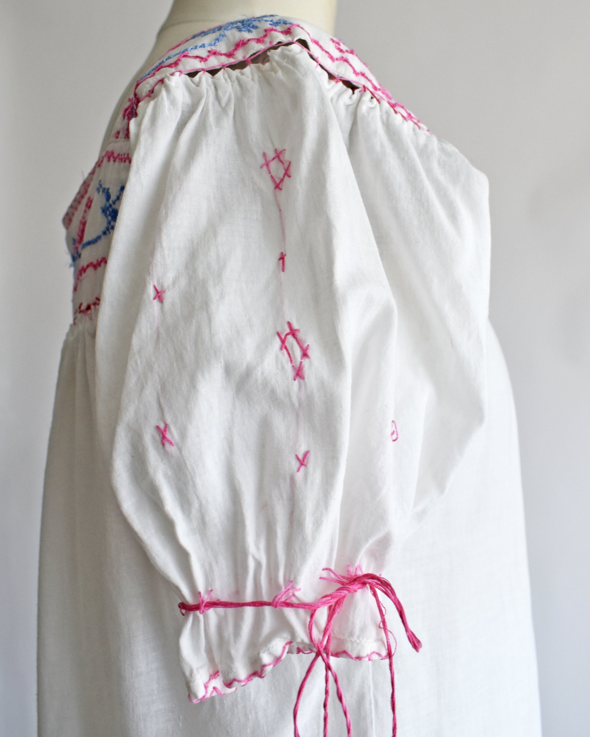 close up of the sleeve on a blouse with pink embroidered details, geometric star patterns, and a pink drawstring tie at the waist, displayed on a mannequin against a plain white background.