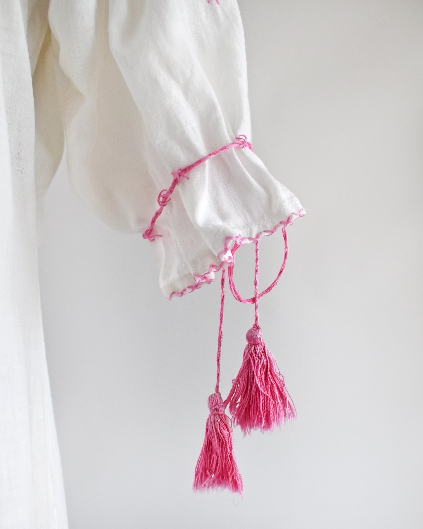 A close-up of a white garment sleeve with bright pink embroidered trim and two hanging tassels, shown against a plain light background.