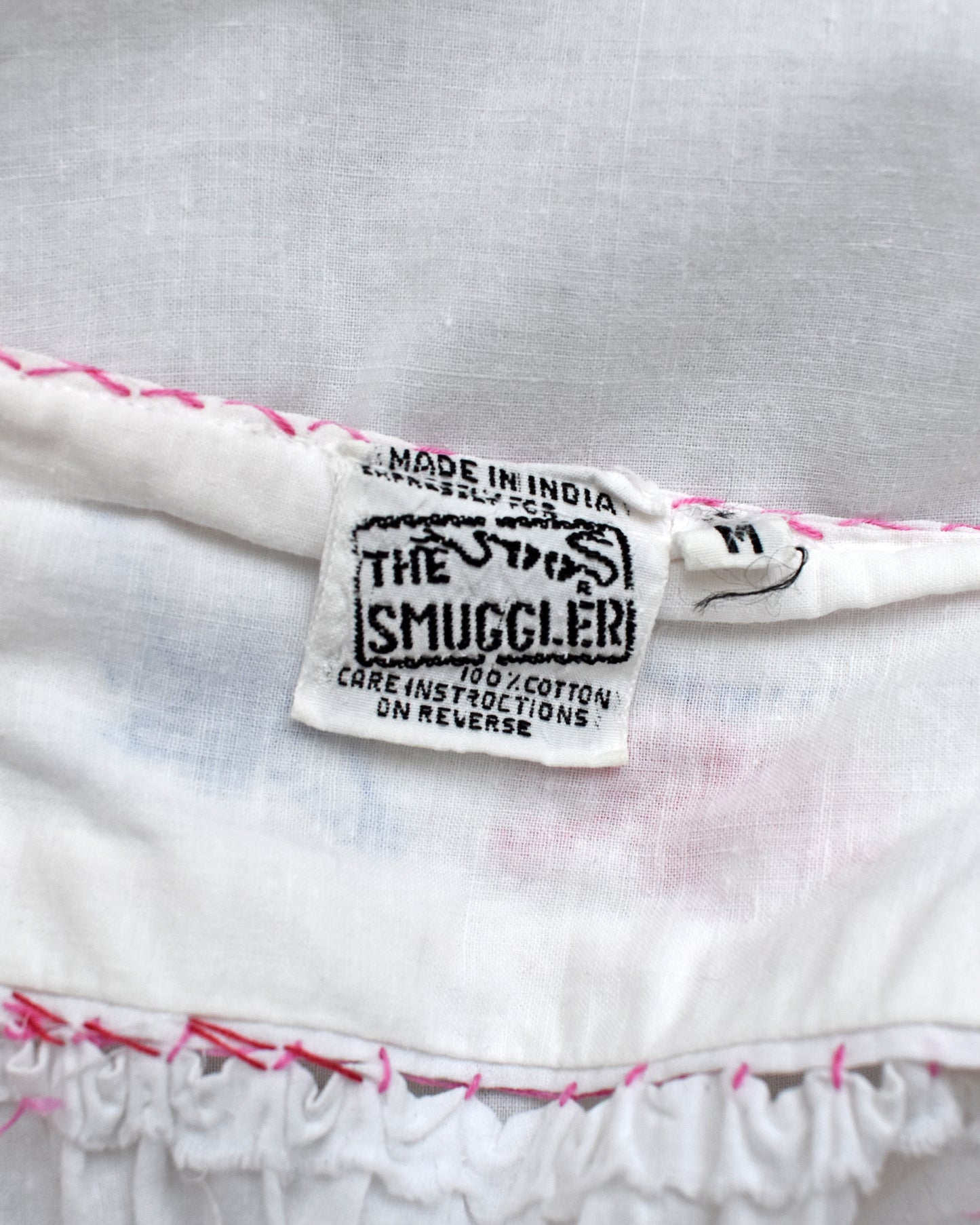 Close-up of a white cotton garment label reading 'THE SMUGGLER' with 'MADE IN INDIA' and care instructions, shown on a white fabric surface with pink stitching.