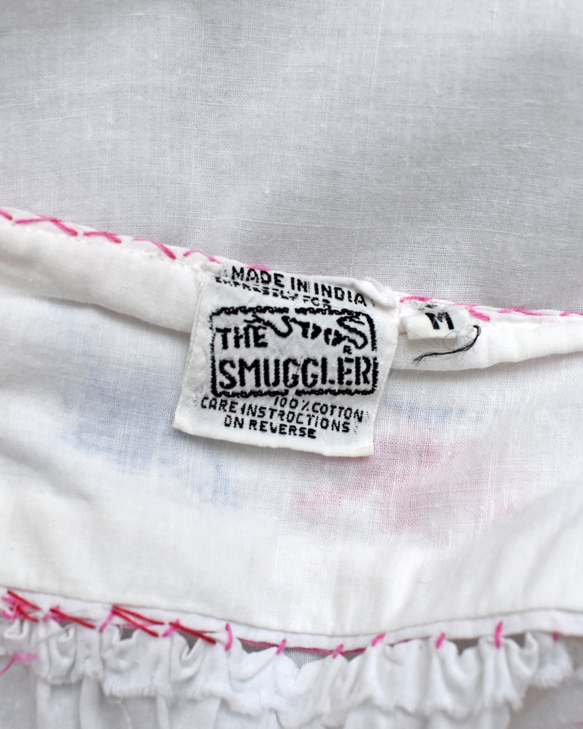 Close-up of a white cotton garment label reading 'THE SMUGGLER' with 'MADE IN INDIA' and care instructions, shown on a white fabric surface with pink stitching.