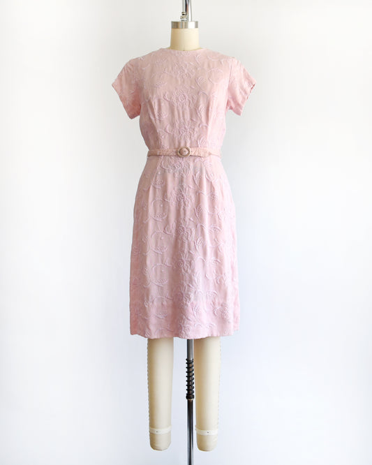A vintage 1950s/1960s light pink short-sleeve dress with an embroidered floral pattern and a matching waist belt, displayed on a cream-colored dress form against a white background.